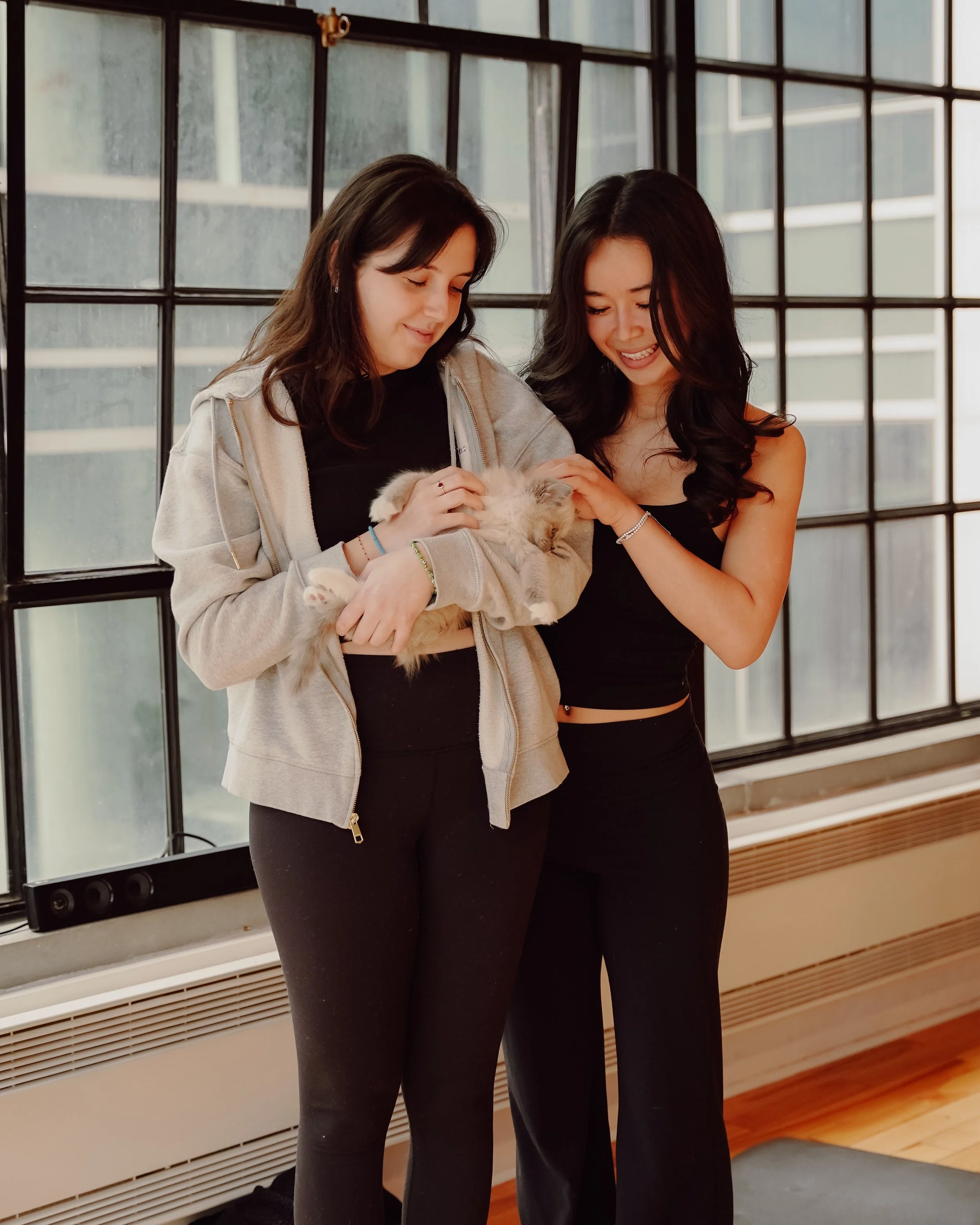 Two women smiling and holding a fluffy kitten inside a room with large windows.