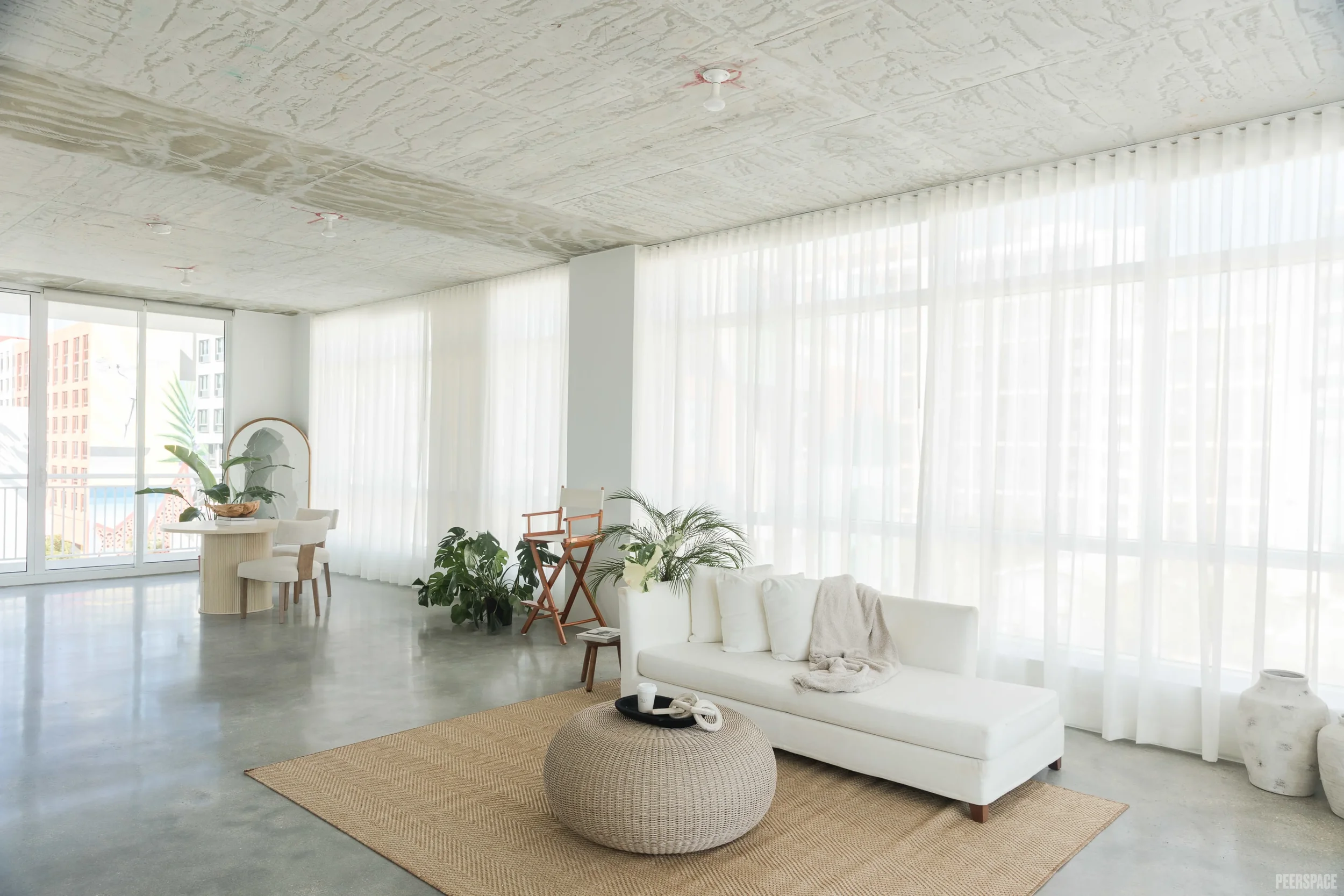 Bright and minimalistic living room with white sofa, round woven coffee table, potted plants, and large windows with sheer curtains.