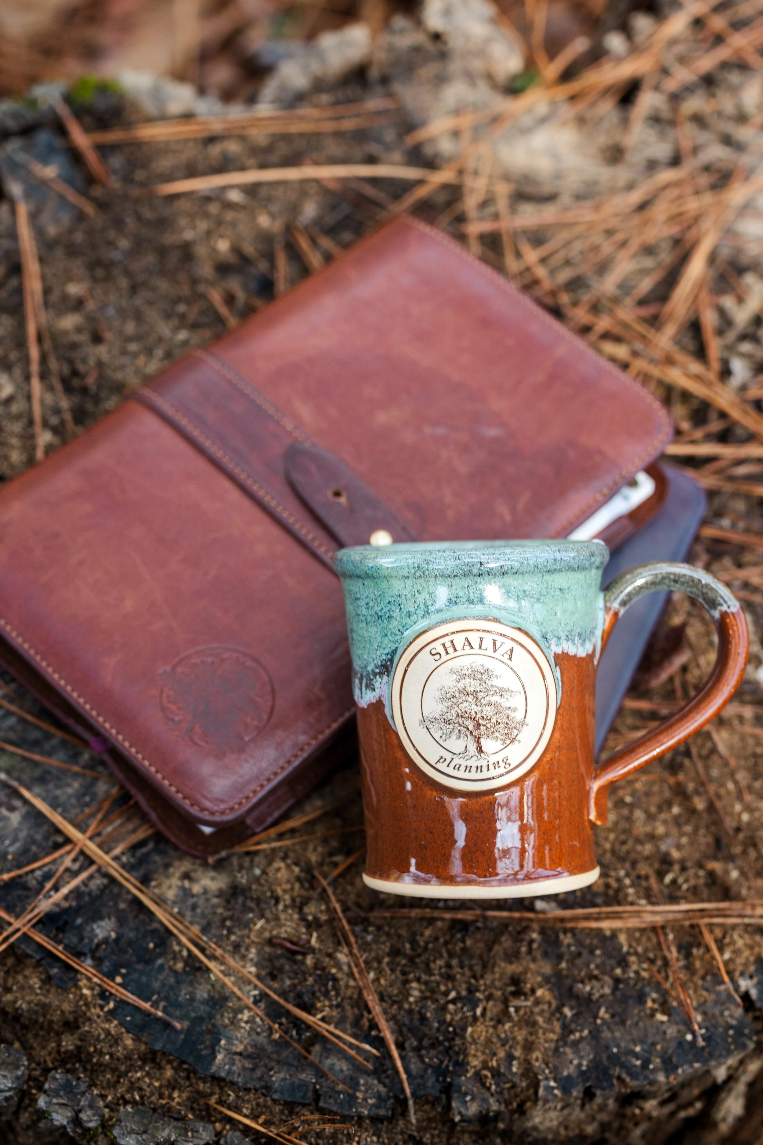 A ceramic mug with a handle, bearing a logo that says 'SHALVA Planning' with a tree illustration, placed on the ground next to a LifePlan Process notebooks and guided reflection materials.
