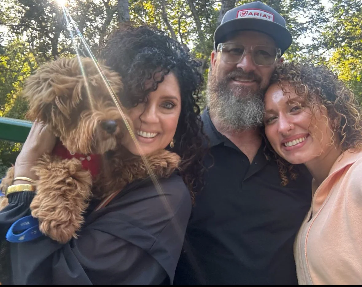 Three people and a dog taking a selfie outdoors with trees in the background. One woman is holding a small brown dog. The man is wearing glasses and a cap. Everyone is smiling.