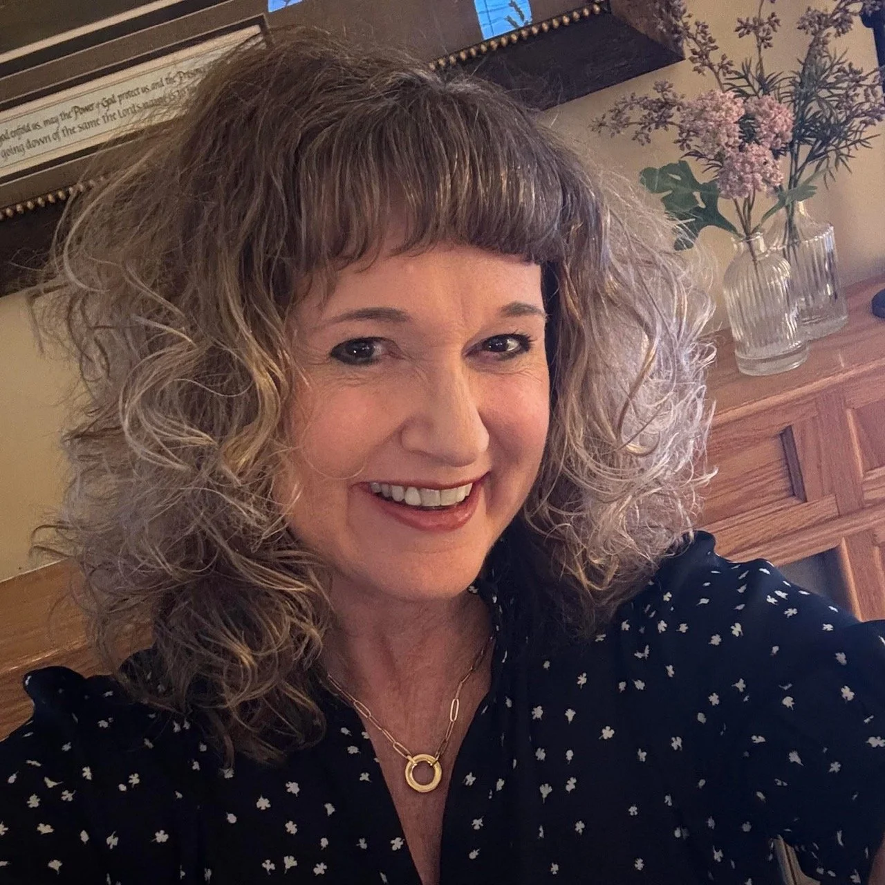 A woman with curly hair and a big smile, wearing a black top with white flower patterns and a gold necklace, taking a selfie in a room with wooden furniture and a vase of pink and purple flowers in the background.