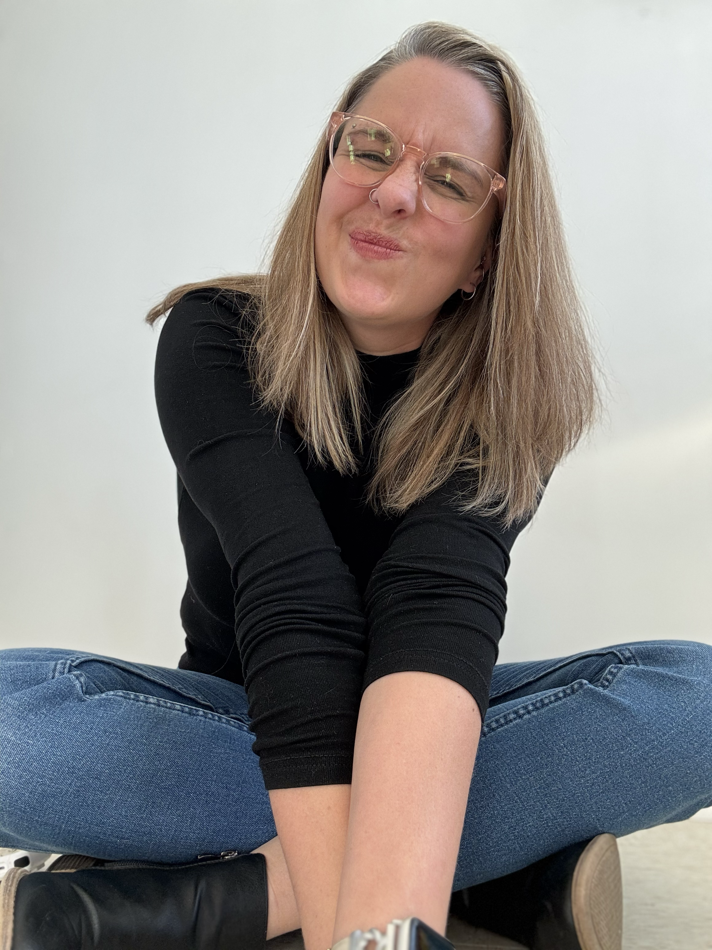 A young woman with glasses, sitting cross-legged, making a funny face with her lips puckered and scrunched eyes, against a plain white background.