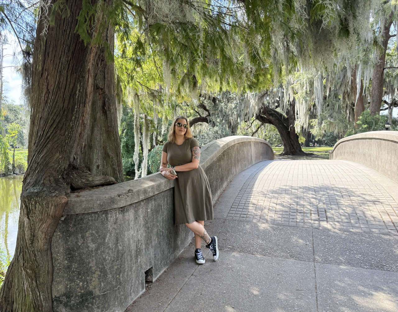 A woman with tattoos wearing sunglasses, a green t-shirt, and a long skirt, standing next to a tree on a stone bridge in a park with hanging moss and large trees.