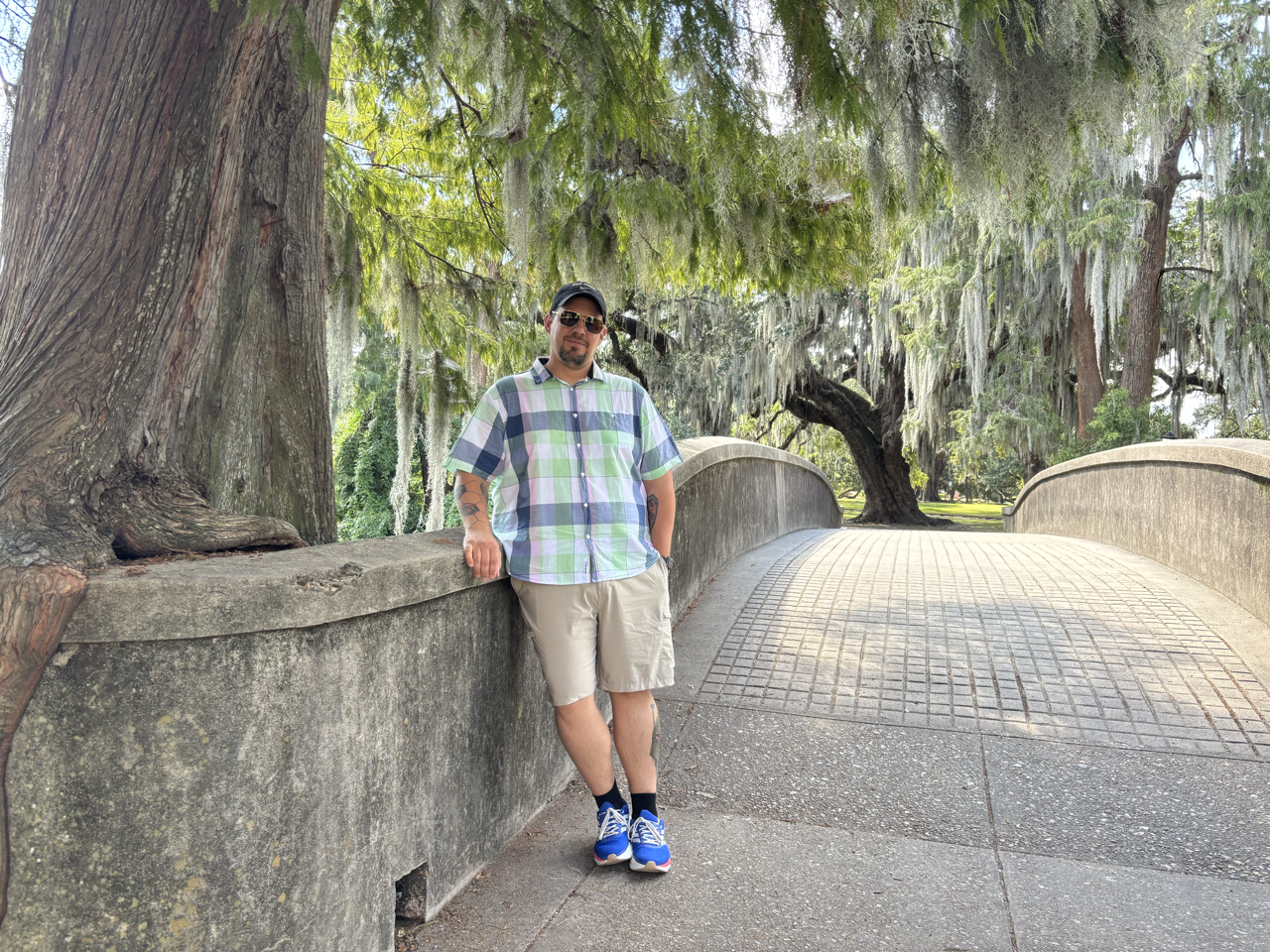 A man standing on a stone bridge with trees and Spanish moss hanging overhead, wearing sunglasses, a plaid short-sleeve shirt, shorts, and blue sneakers.