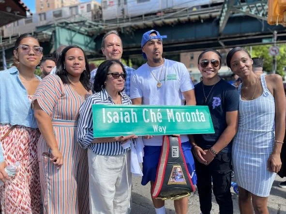 Seven people standing outdoors under a bridge, holding a street sign that reads 'Isaiah Ché Moronta Way.' They are smiling and honoring Isaiah in Washington Heights. Non-profit inspired by the bravery of Isaiah Che Moronta.