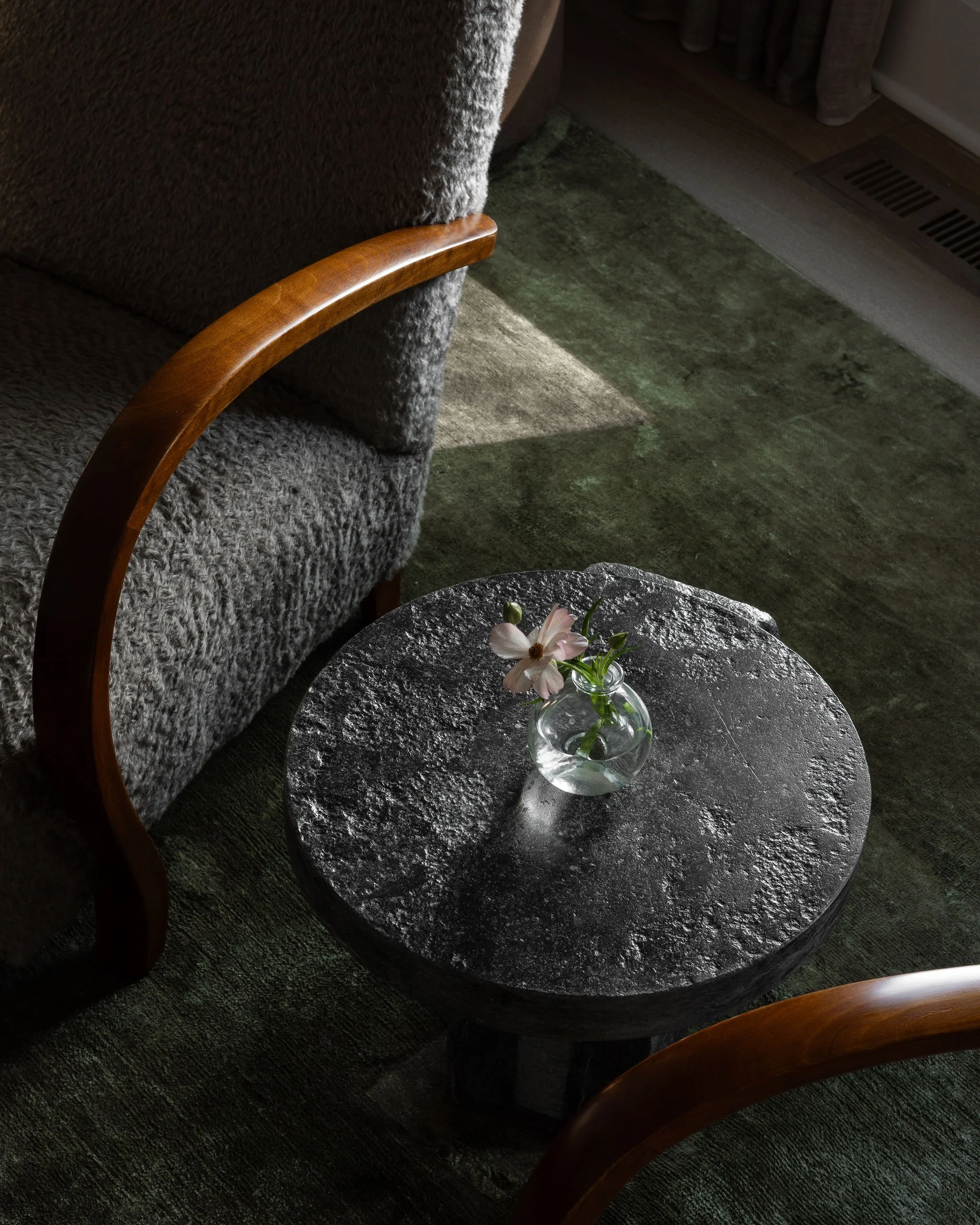 Vignette with a sculptural stone table by Maverick Lee between vintage cabinetmaker chairs, set on a green linen rug with soft light on a single pink flower.