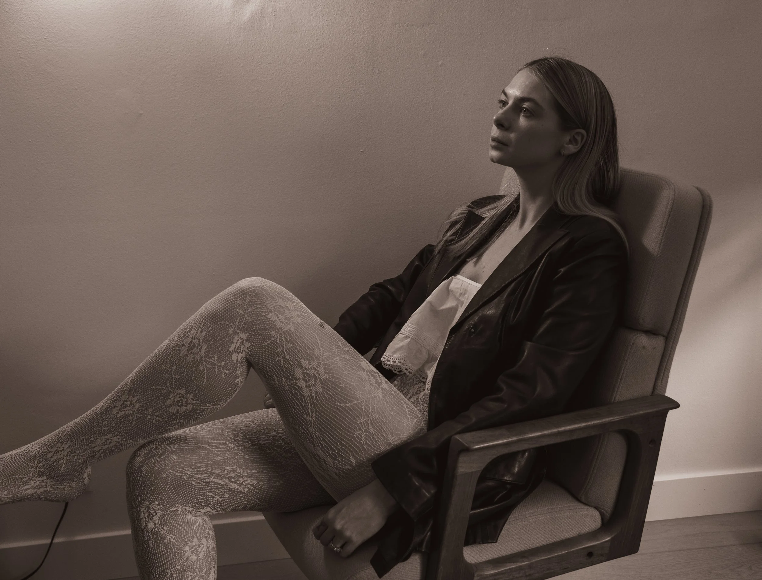 A woman sitting in a wooden and fabric chair against a plain wall, wearing an upcycled black leather jacket and lace-patterned tights, gazing slightly upward with a contemplative expression.