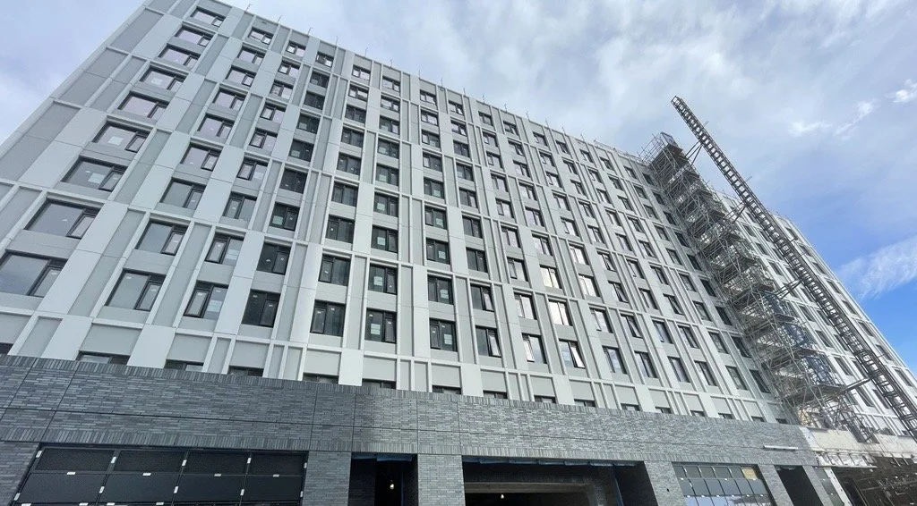 View of a modern multi-story building under construction with scaffolding and a tower crane on the right side, sky in the background.