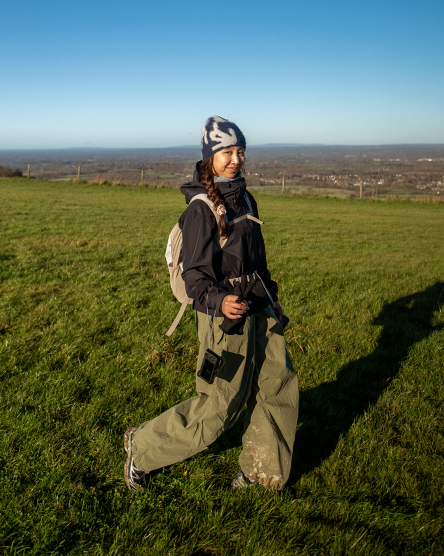 Young woman with a backpack and winter hat walking on a grassy hilltop under a blue sky.