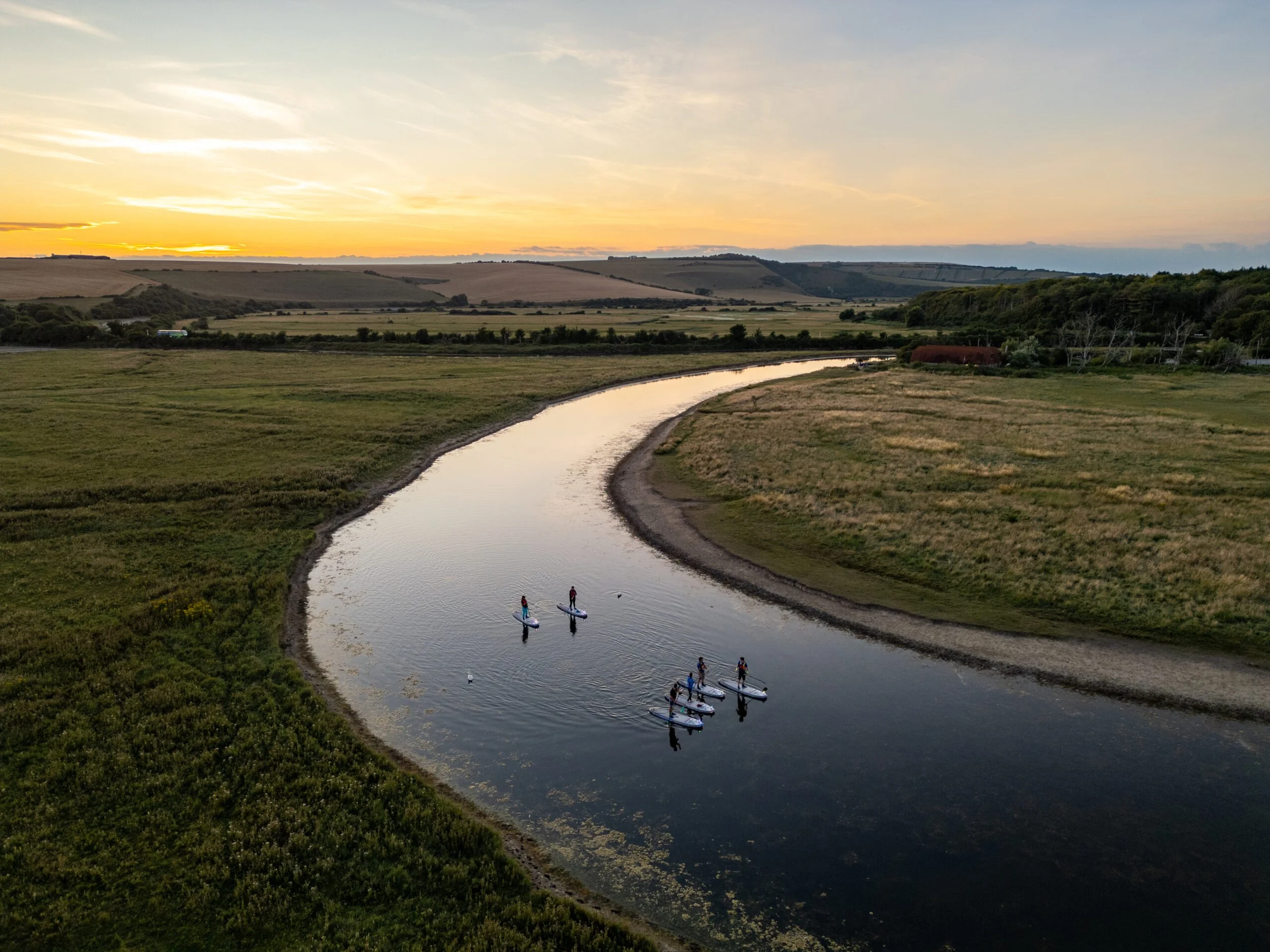 A group of people are paddleboarding on a winding river at sunset, surrounded by green fields and distant hills.