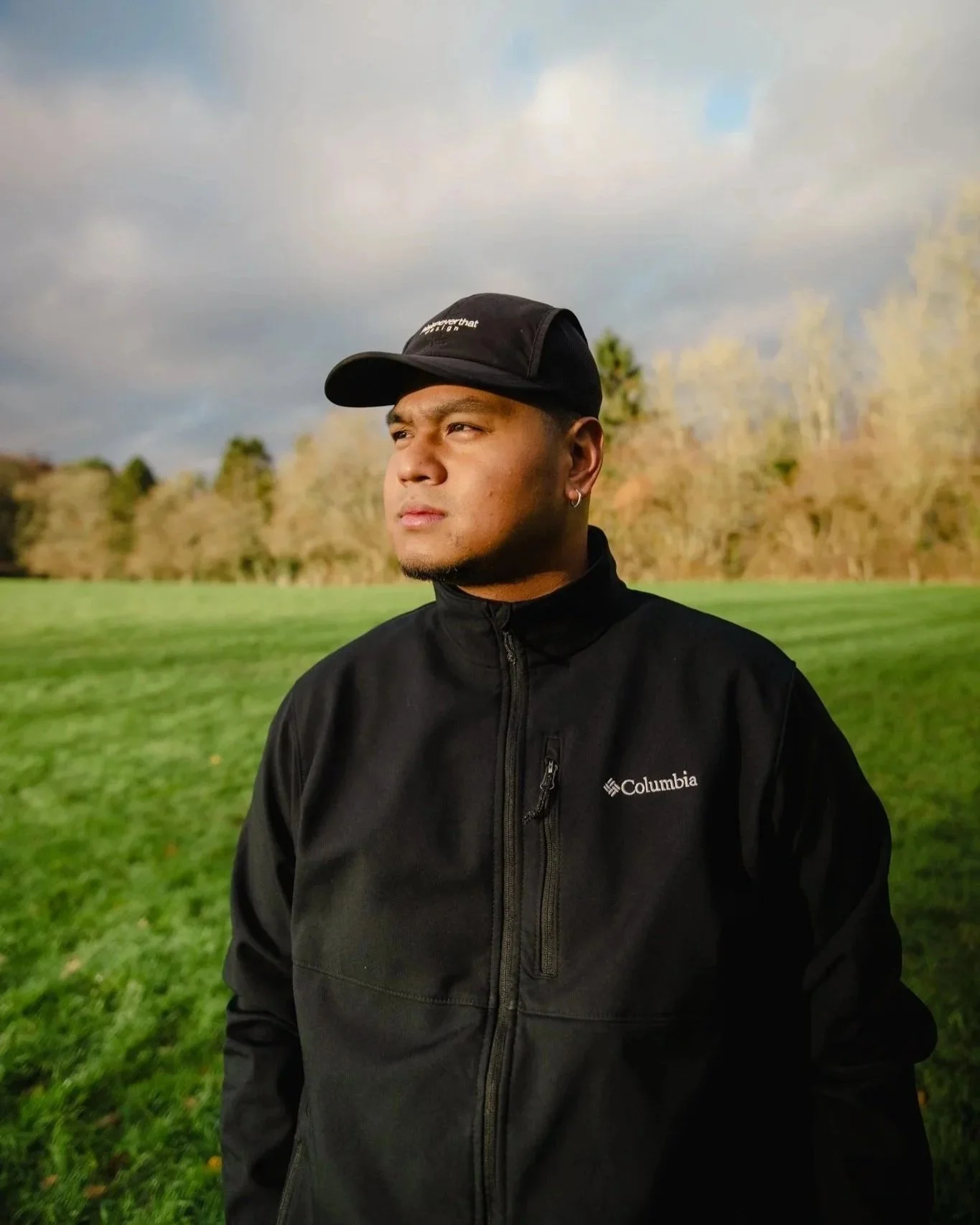 A man wearing a black Columbia jacket and baseball cap, standing outdoors in a grassy field with trees and a cloudy sky in the background.