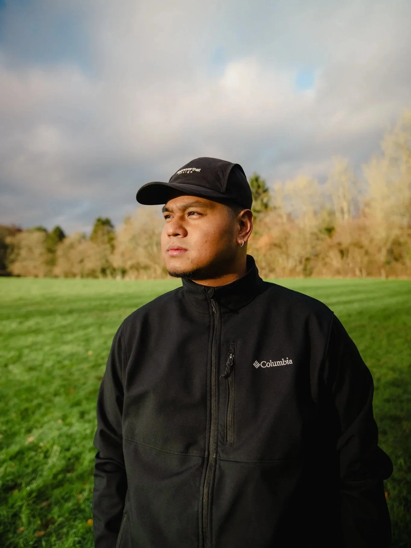 Young man standing outdoors in a grassy field with trees in the background, wearing a black Columbia jacket and black cap, looking to the left.