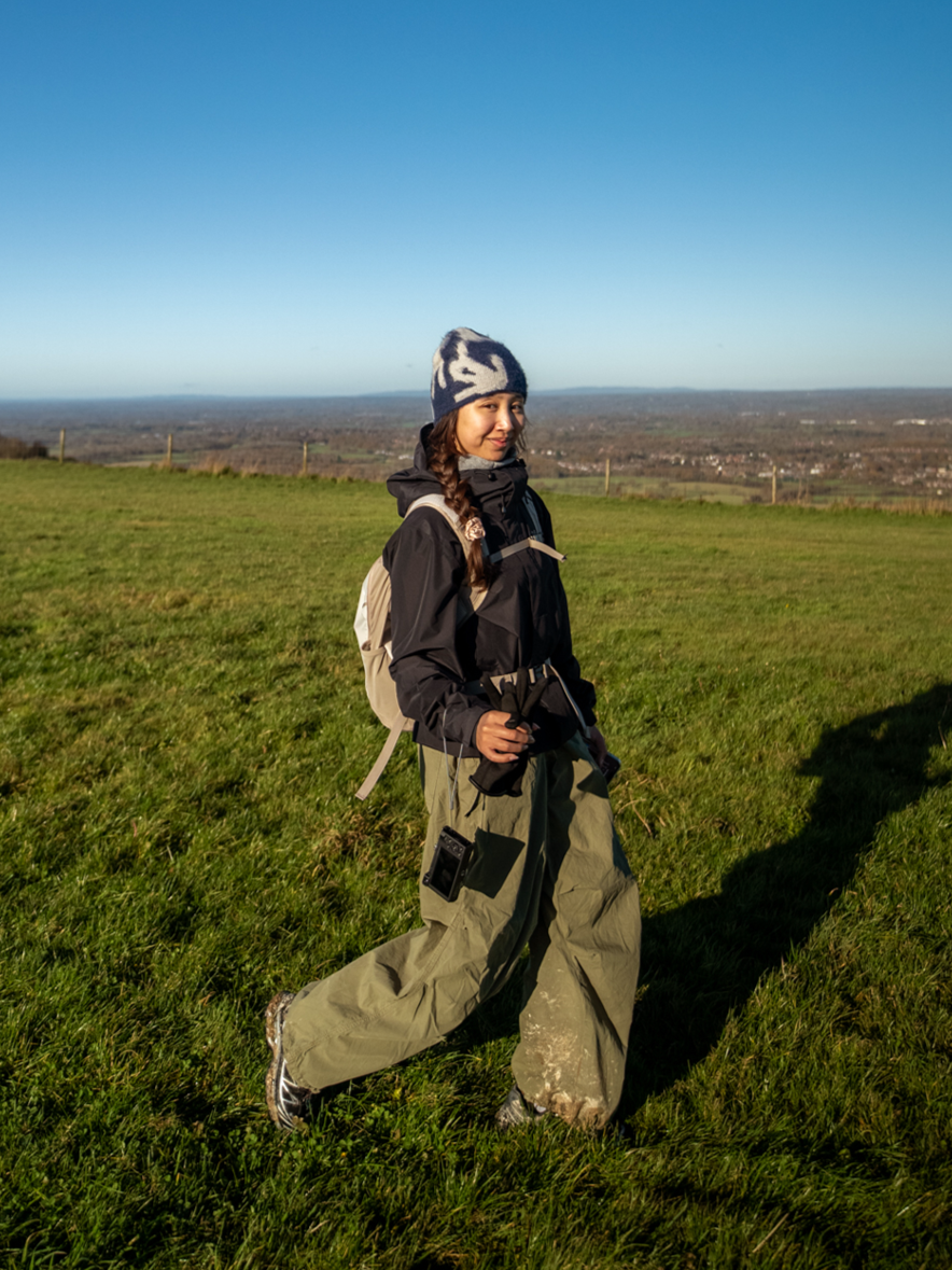 A woman hiking on a grassy hill, dressed in outdoor clothing including a beanie, jacket, and bag, with a scenic view of the countryside in the background.