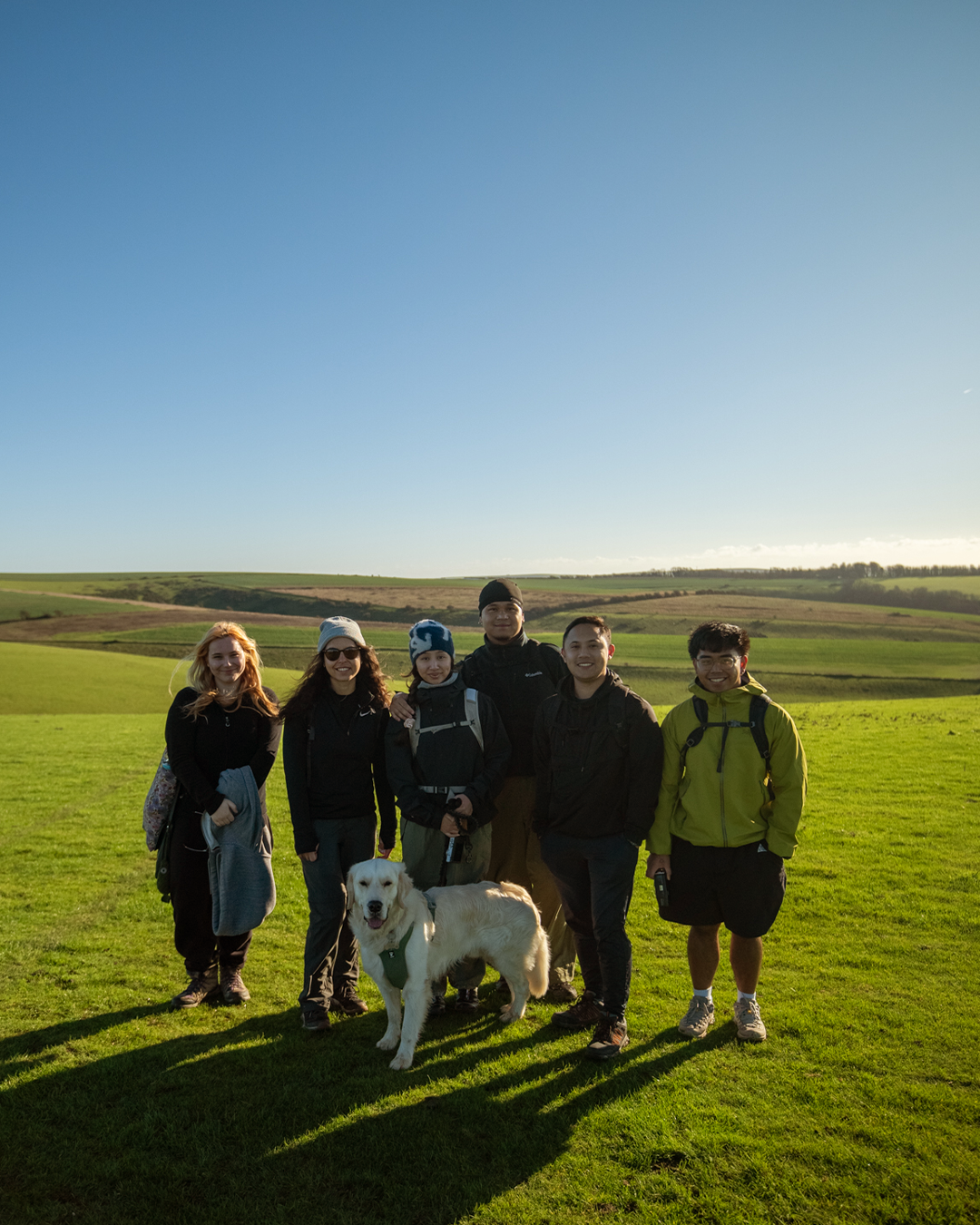 Group of six people and a dog standing outdoors on grassy field under clear blue sky.