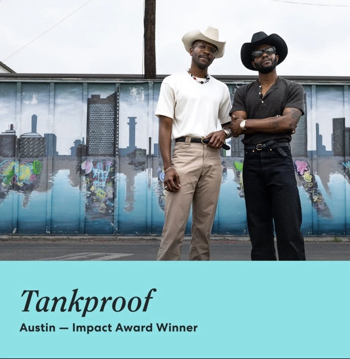 Two men standing in front of a graffiti mural of a city skyline in Austin. One in beige pants and a white shirt, the other in black pants and a black shirt. Both wearing hats and sunglasses.