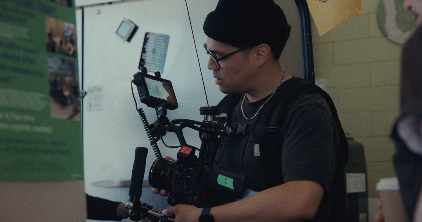 A man with black hair, glasses, a black beanie, and a silver chain, holding a professional camera rig and looking at a monitor, standing indoors near a white wall and a green bulletin board.