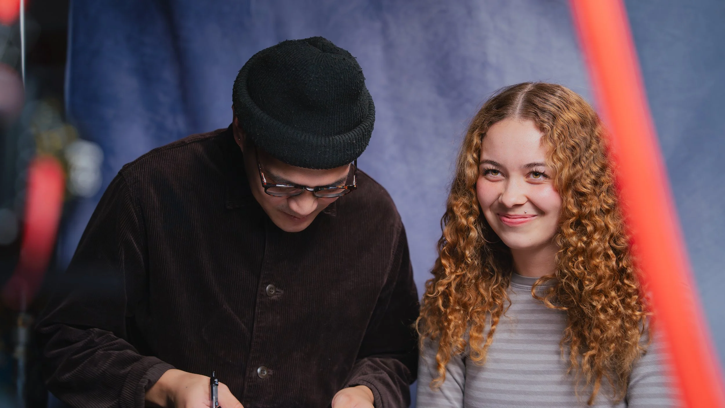 A young woman with curly red hair and a lip piercing smiling, and a man wearing glasses and a black beanie looking down, in an indoor setting with a dark blue background.