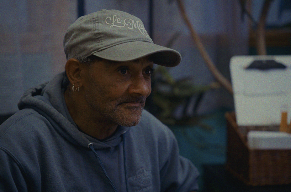 A man wearing a beige baseball cap and a gray hoodie sitting indoors with a focused expression.