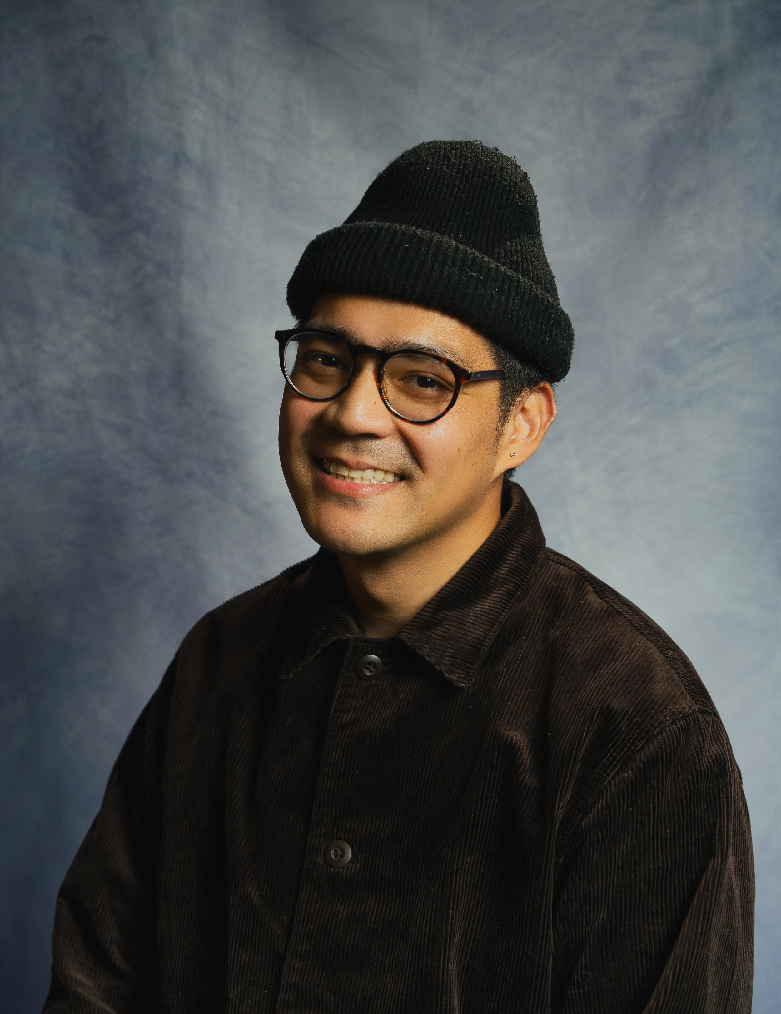 A young man with glasses, wearing a black beanie and a dark brown corduroy shirt, smiling at the camera against a textured blue-gray background.