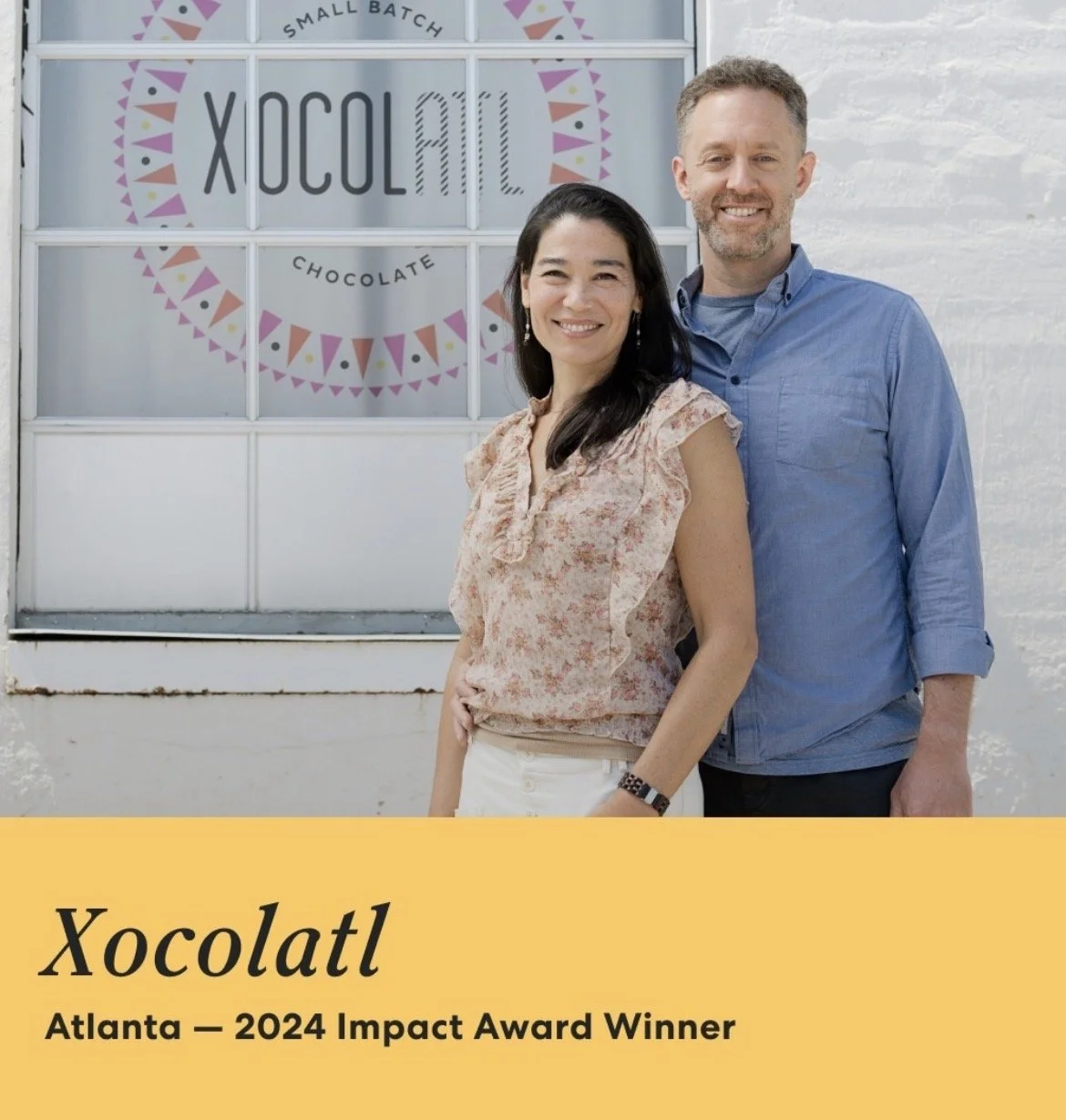 A man and woman standing outside a small bakery named Xocotatl, which won the 2024 Impact Award in Atlanta. The woman is wearing a floral blouse and the man is wearing a blue shirt, both smiling.