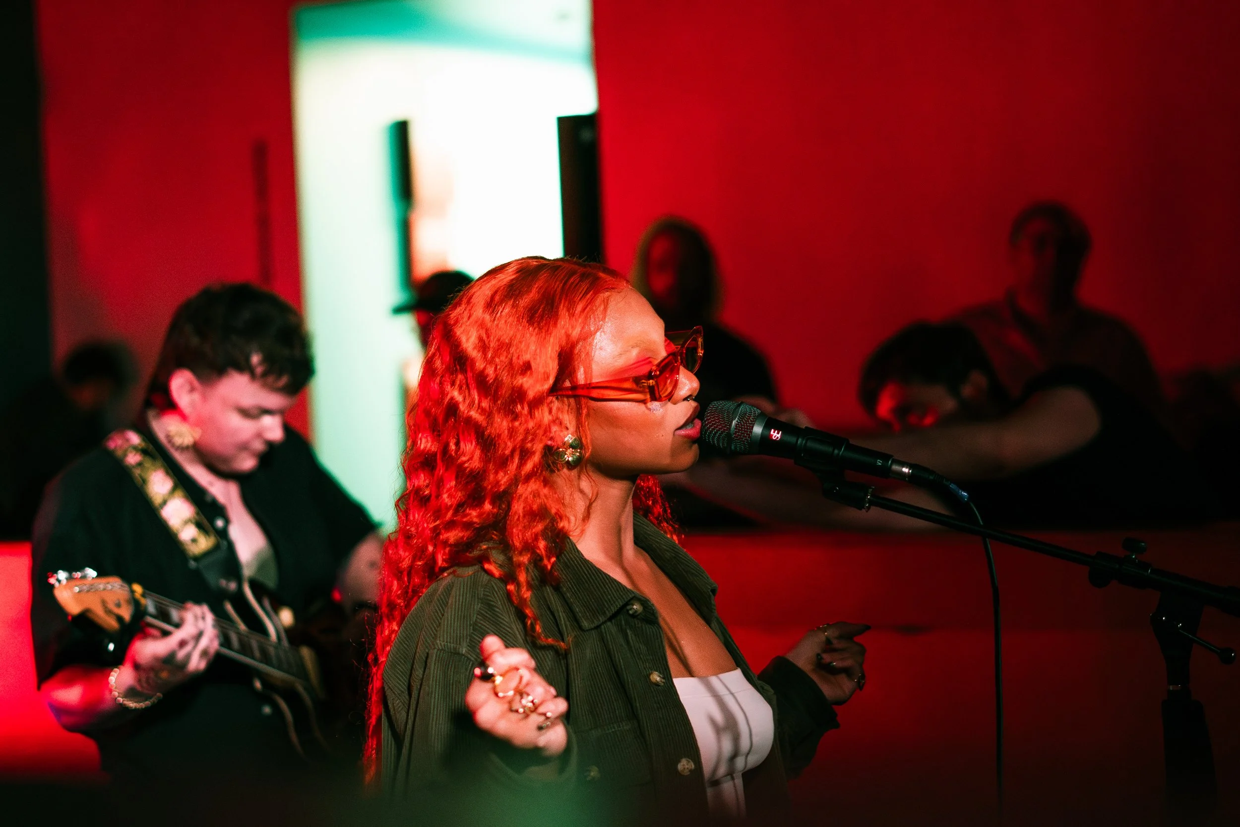 A woman with long red curly hair, wearing glasses and a green jacket, singing into a microphone during a performance, accompanied by a guitarist in a black outfit in a dimly lit room with red and green lighting.
