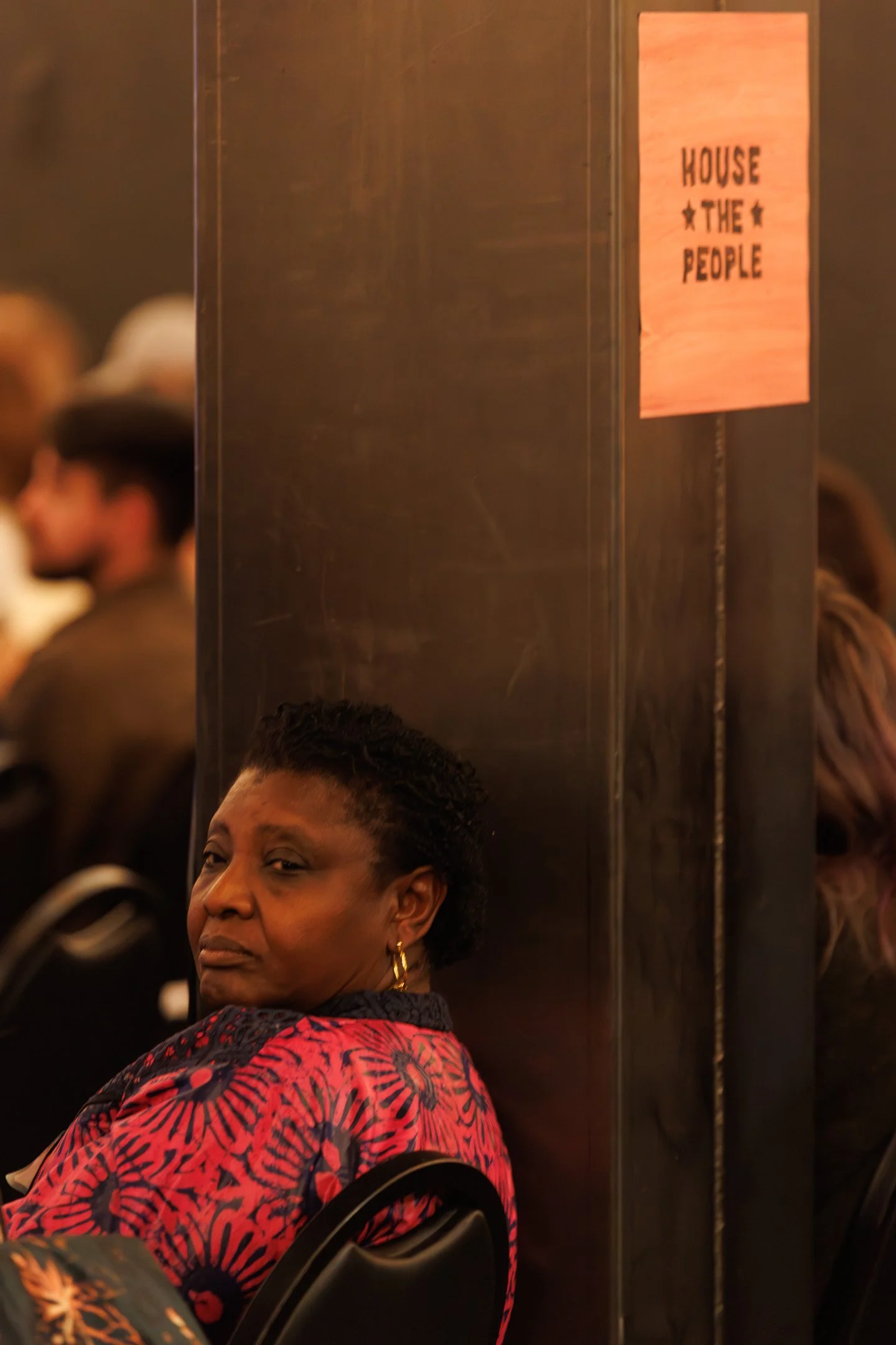 A woman sitting on a chair, looking at the camera, with a dark wall behind her. There is a sign on the wall that reads 'HOUSE OF THE PEOPLE'.