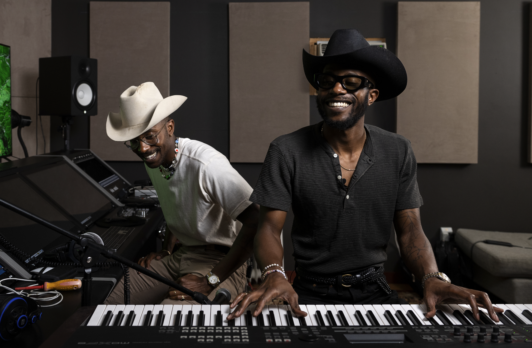 Two men in a recording studio, one playing a keyboard and the other wearing a cowboy hat, both smiling