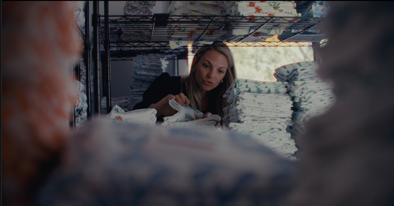Woman sorting folded cloths or linens on shelves inside a room, seen from a low angle through blurred foreground objects, with a window in the background.