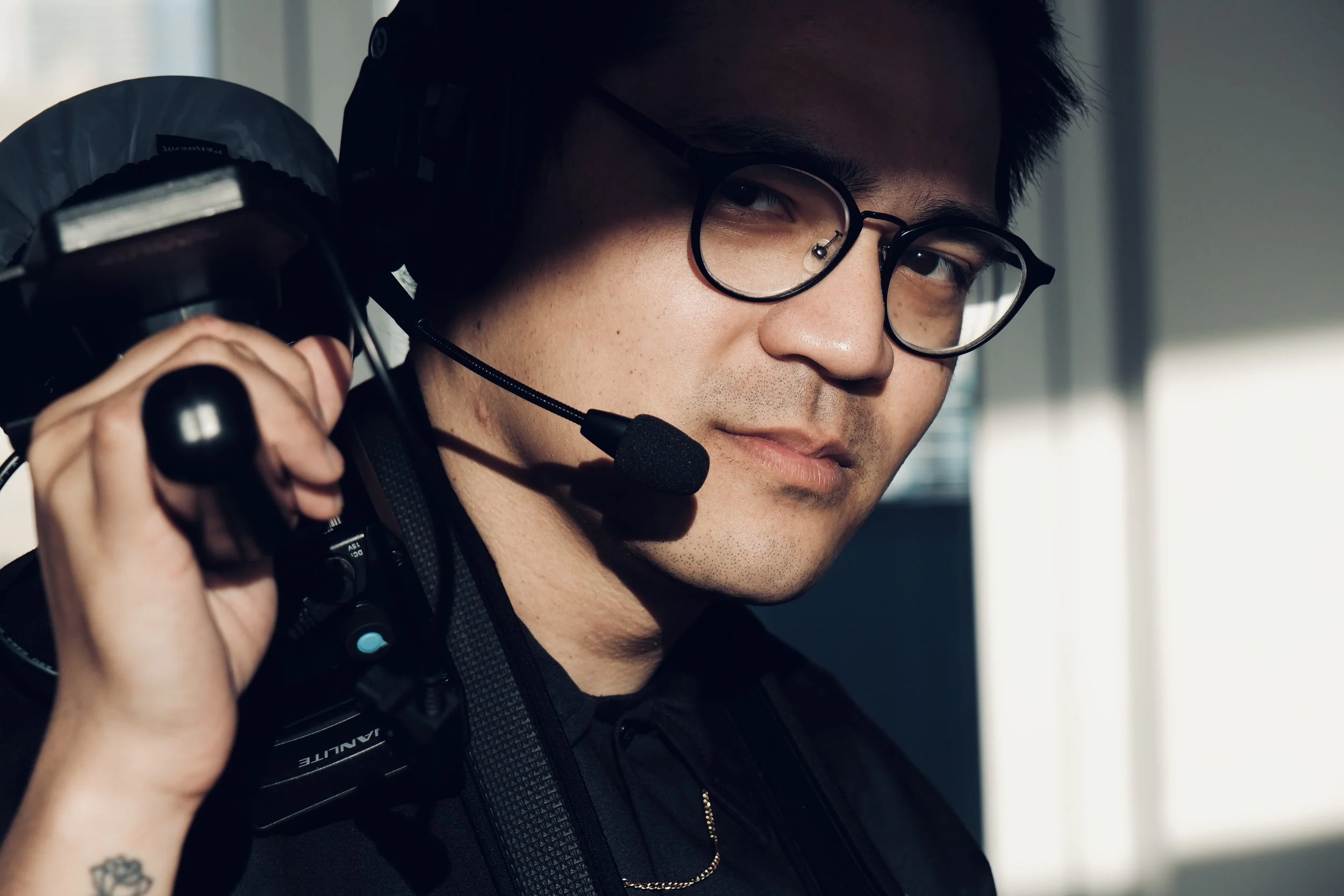 Close-up of a man wearing glasses and a headset, smiling and holding a shoulder strap, with a window in the background.