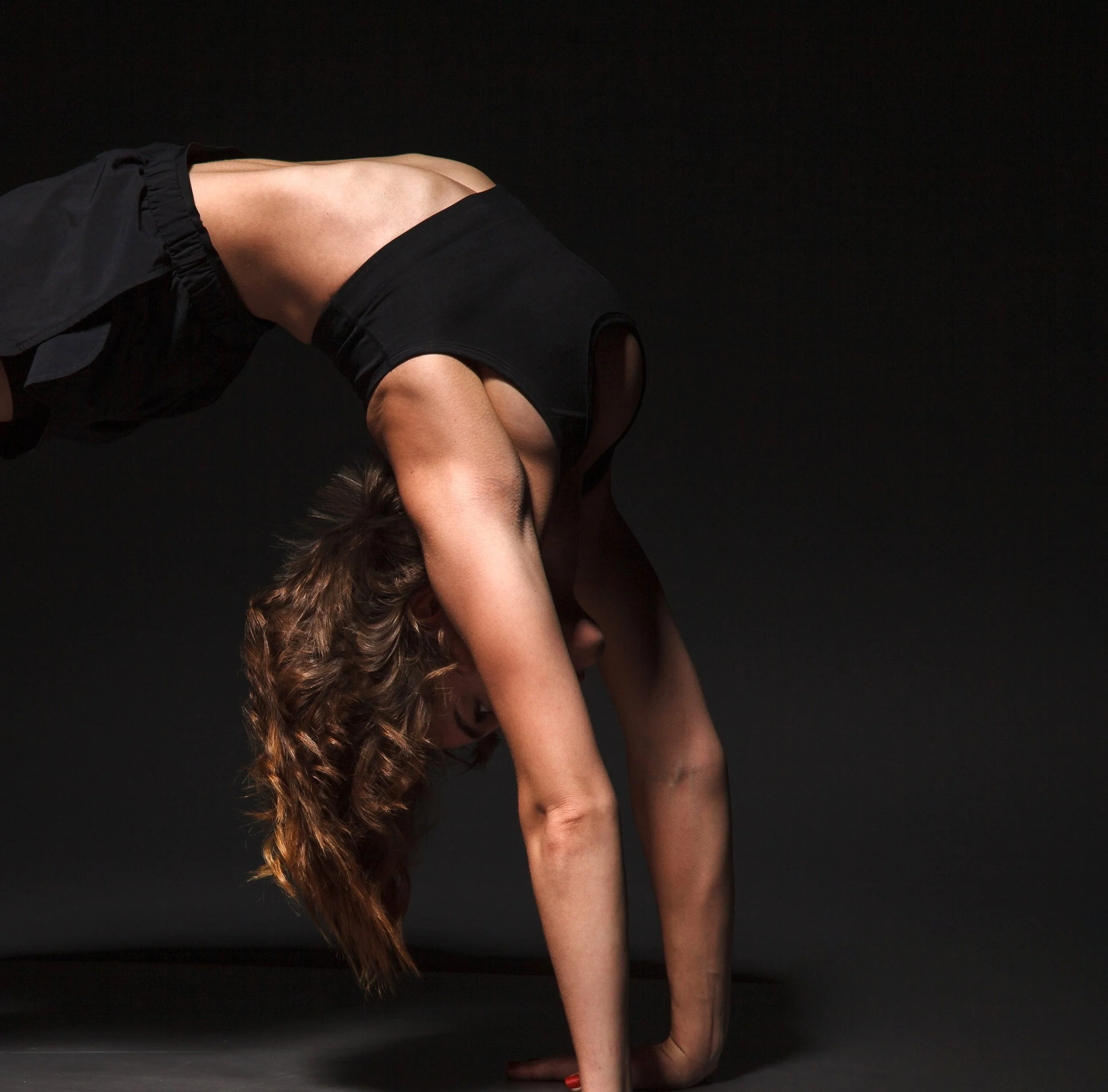 A woman with long, curly hair in a black sleeveless top and black pants is on her hands and knees against a black background, looking downward.