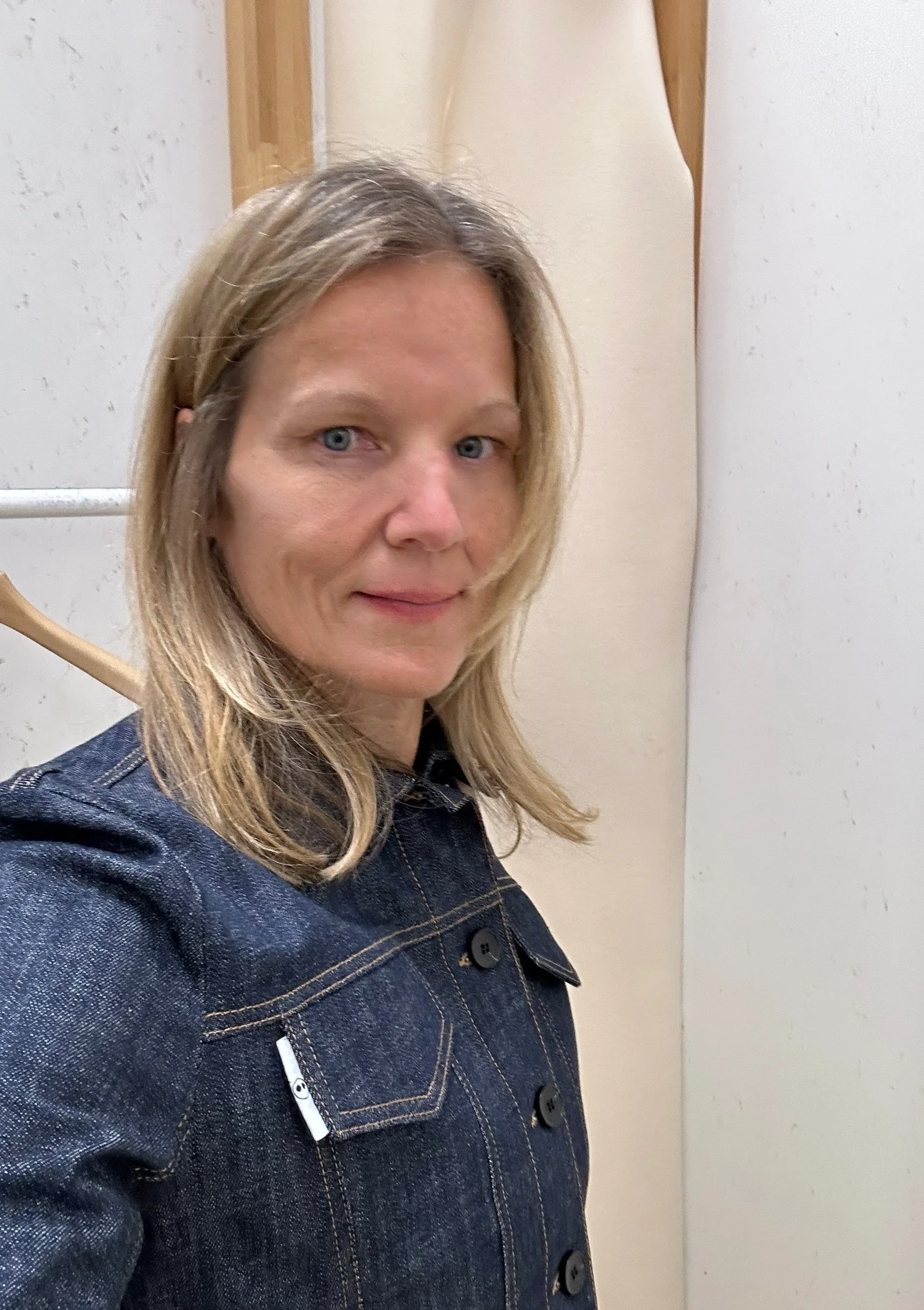 Close-up of a woman with blonde hair, smiling, wearing a denim jacket, standing in front of a plain wall and wooden door.