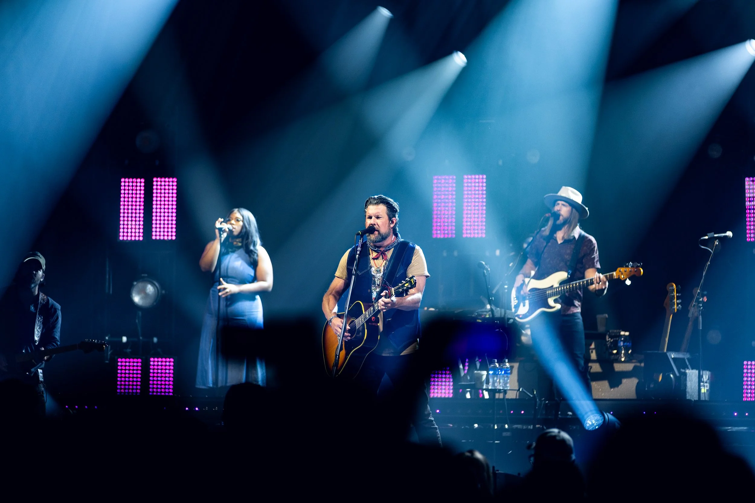 Band performing on stage with bright lights and purple LED screens in the background, including a male musician with a guitar, a female singer, and a bassist with a hat.