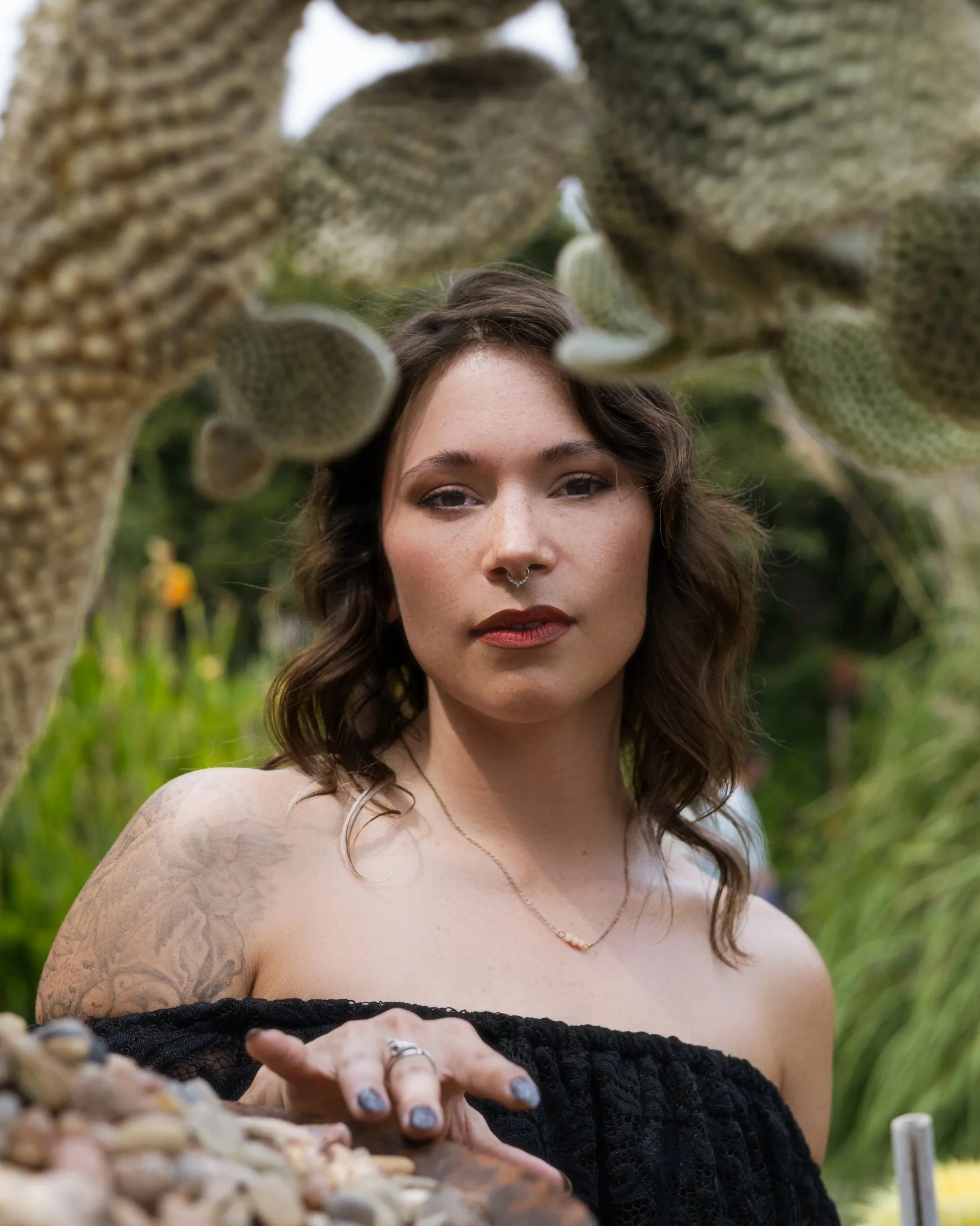 A woman with wavy brown hair, wearing a black off-shoulder top, standing outdoors in a lush green environment. She has tattoos on her left arm, a septum piercing, and is wearing makeup with red lipstick and a necklace. Her hand is extended toward the