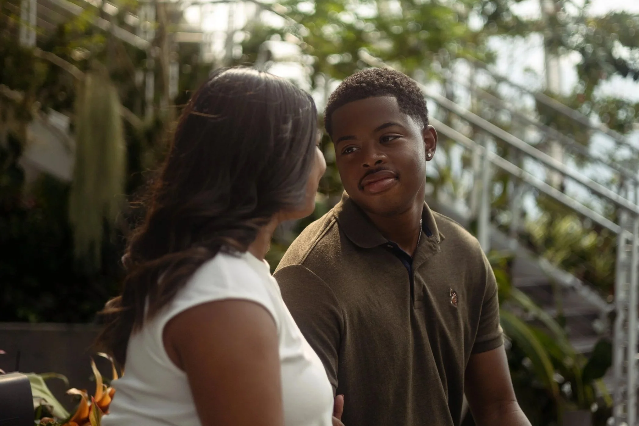 A woman and a young man having a conversation outdoors, with plants and a staircase in the background.
