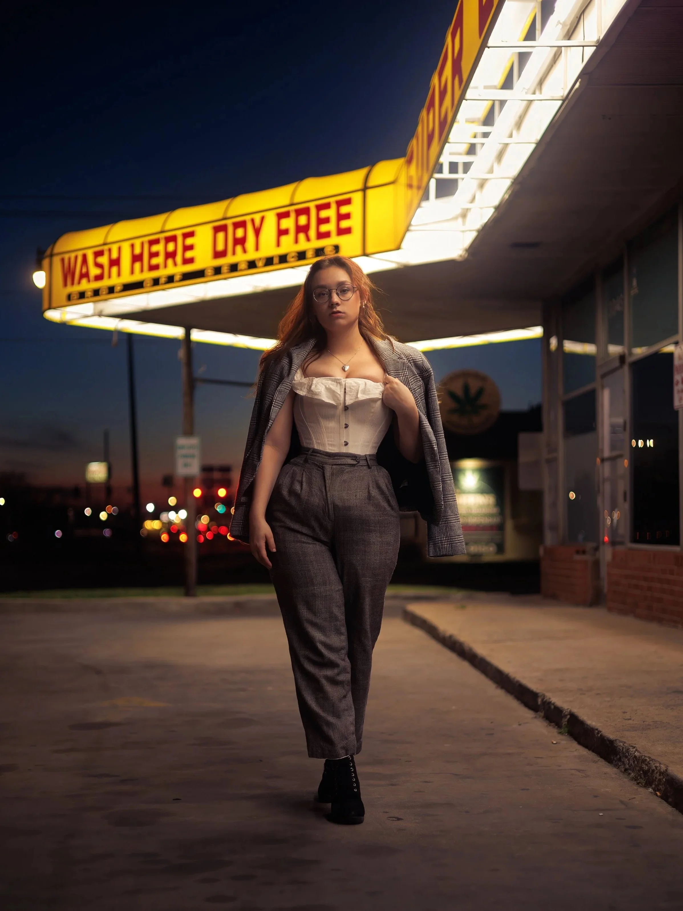 A woman wearing glasses, a white corset top, plaid trousers, and a blazer draped over her shoulders walking outside at night near a brightly lit business with a sign that reads 'Wash Here Dry Free'.
