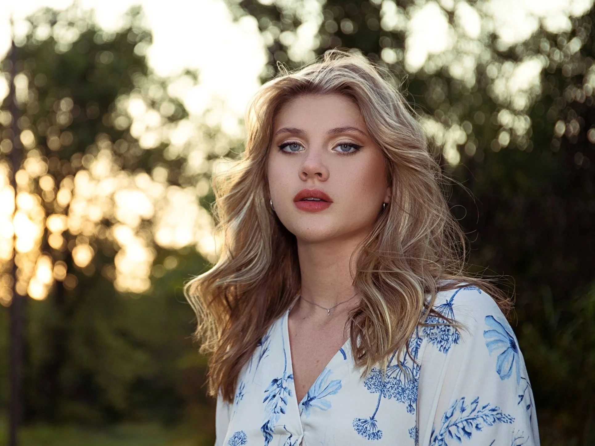 A young woman with wavy blonde hair and blue eyes standing outdoors during sunset, wearing a white blouse with blue floral patterns.