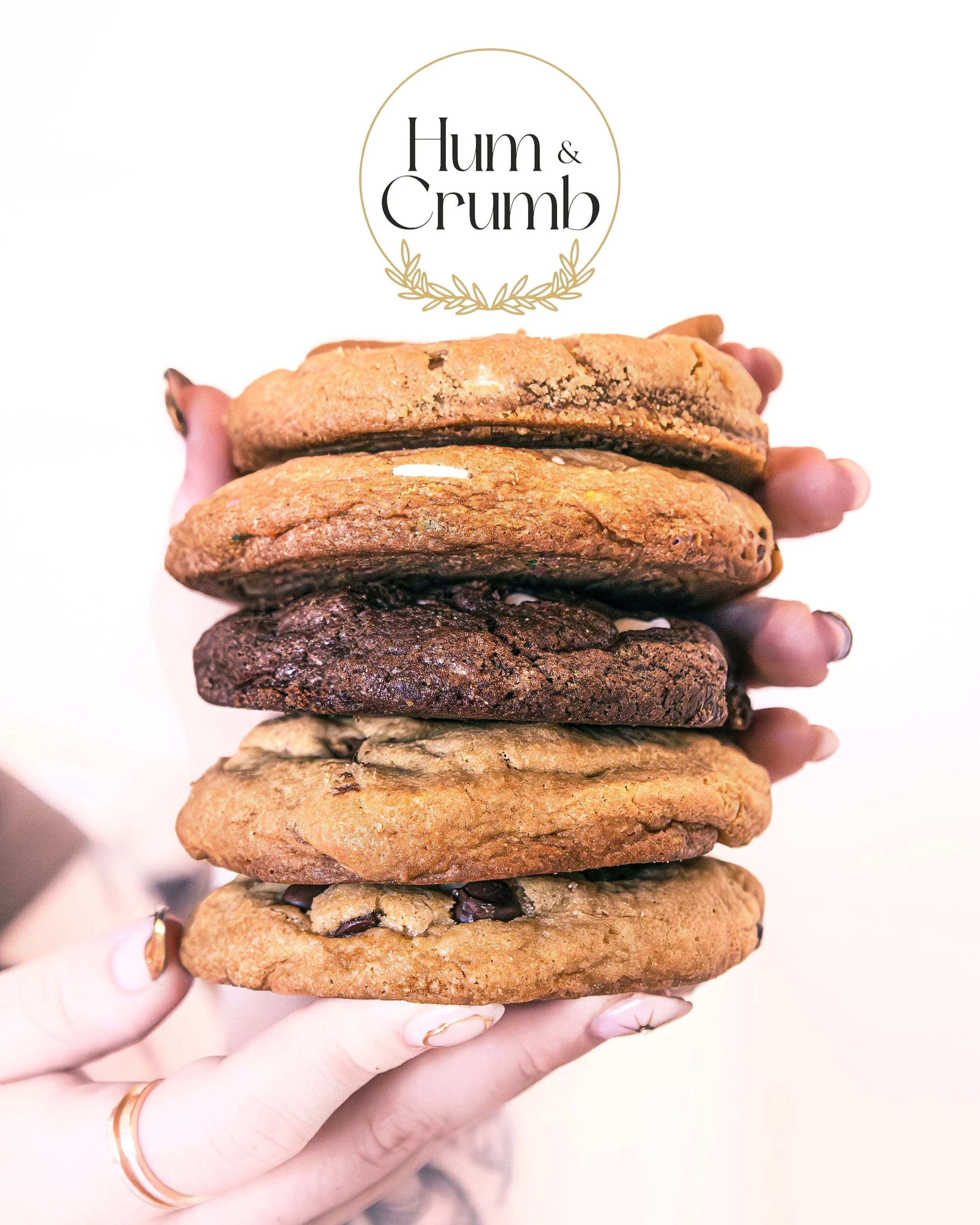 Hand holding a stack of six large cookies, with a logo that says "Hum & Crumb" above the cookies.