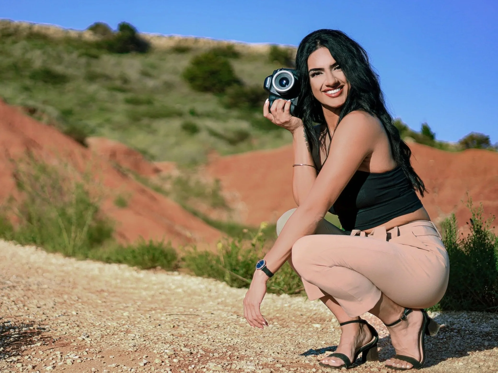 A woman with long black hair squatting outdoors, holding a camera, with red rock formations and green hills in the background, under a clear blue sky.