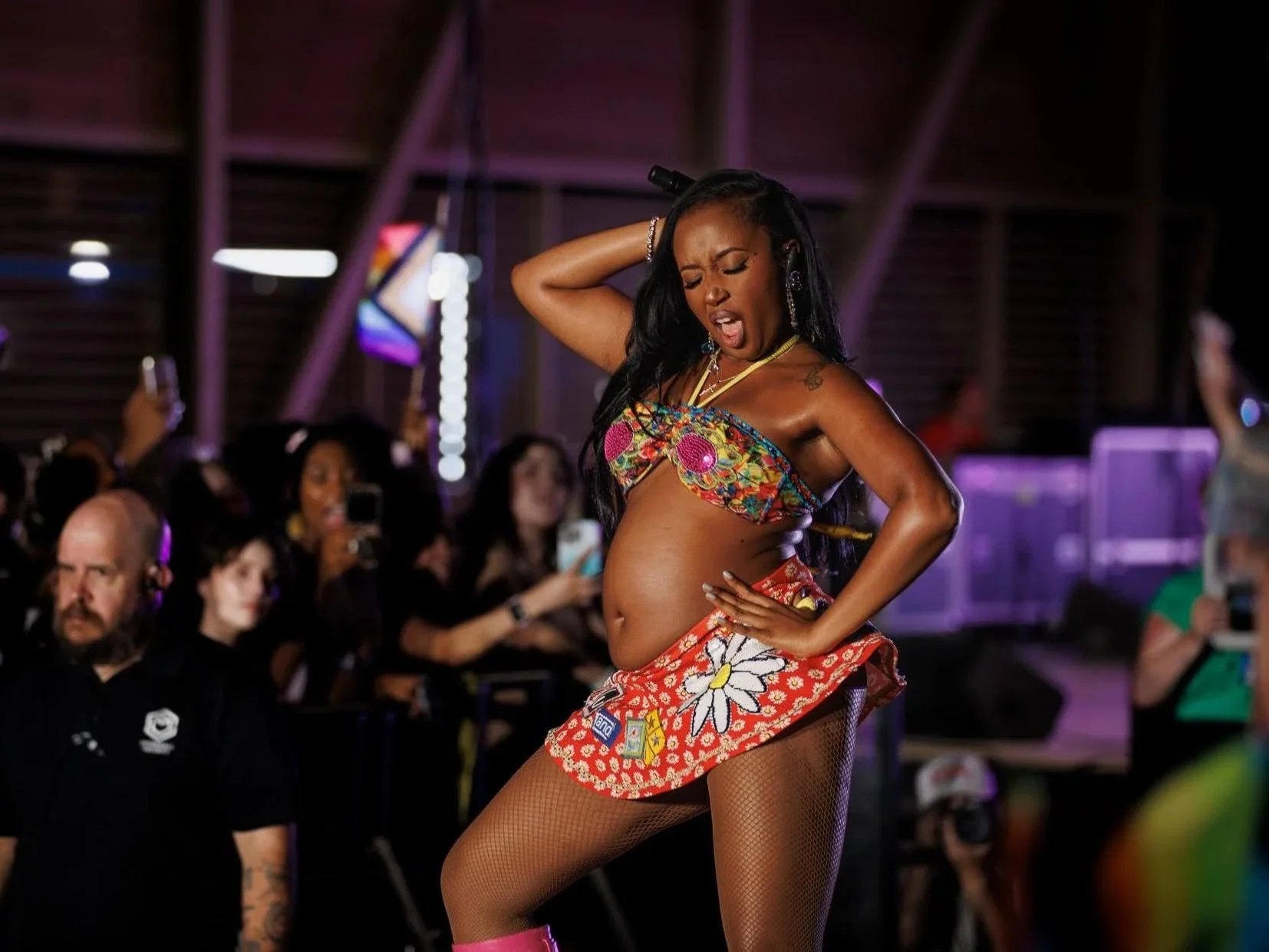A woman dancing on stage, wearing colorful clothing, with a crowd of people taking photos and cheering in the background.