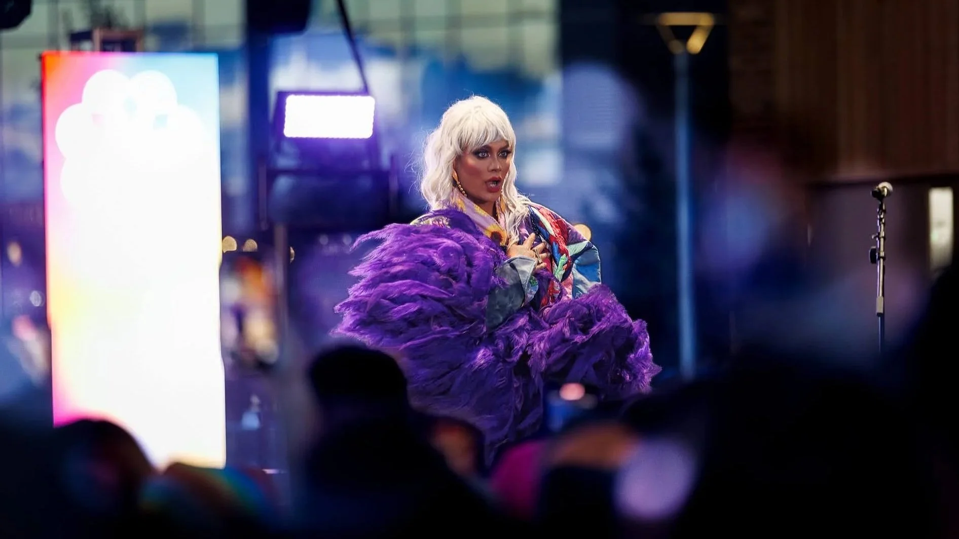 A person with platinum blonde hair performing on stage, wearing a colorful jacket and holding a large purple feathered fan, with a microphone stand nearby and bright stage lights in the background.