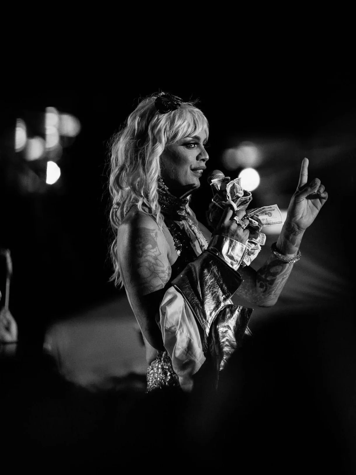 Black and white photo of a female performer with blonde hair, holding a microphone wrapped in dollar bills, raising one finger, on a dark stage with blurred lights in the background.