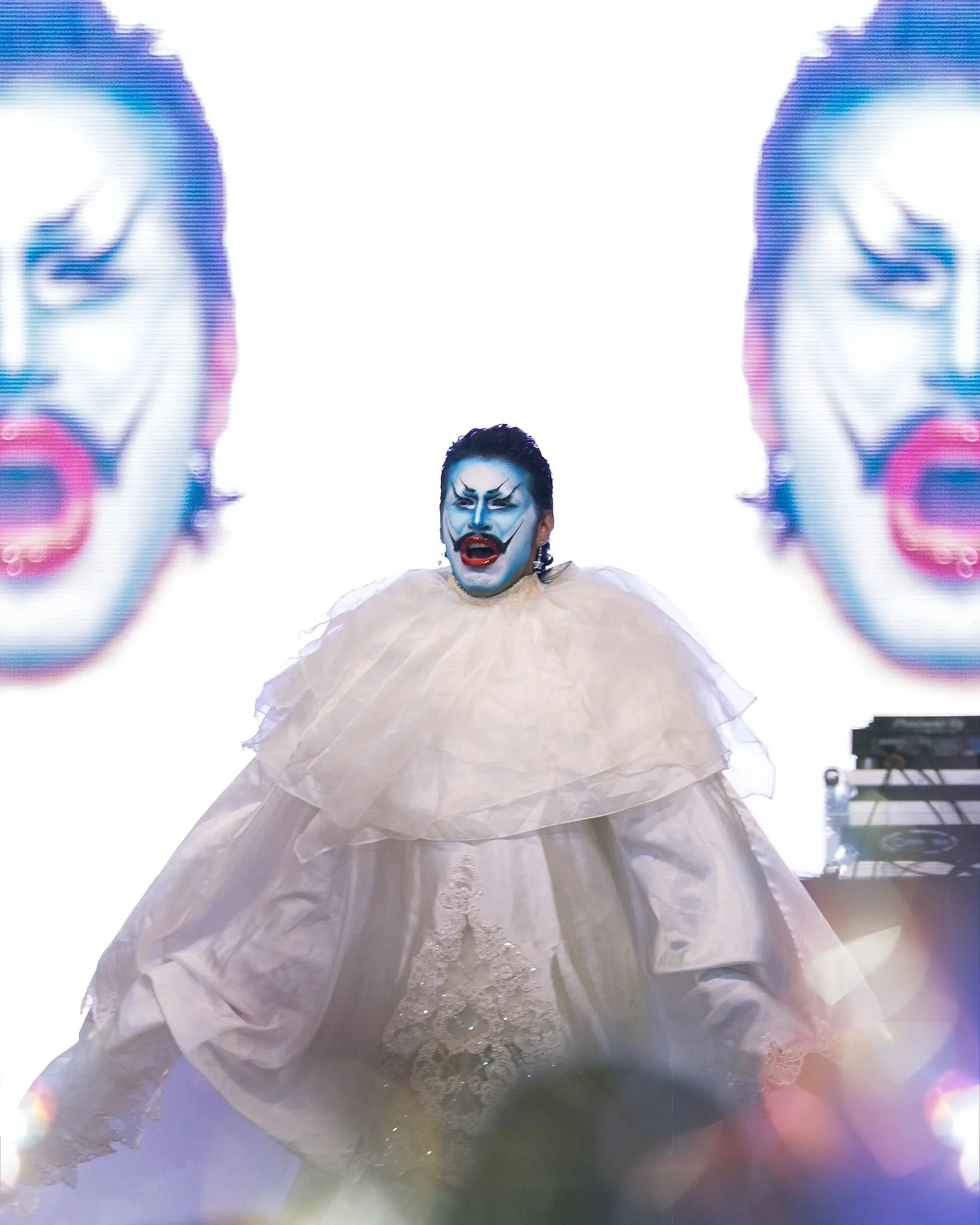 A performer with blue face makeup and dark hair styled back, wearing a dramatic, layered white gown with lace and tulle, singing on stage with bright white background and two large, distorted screens of a clown face projected behind, along with a DJ 