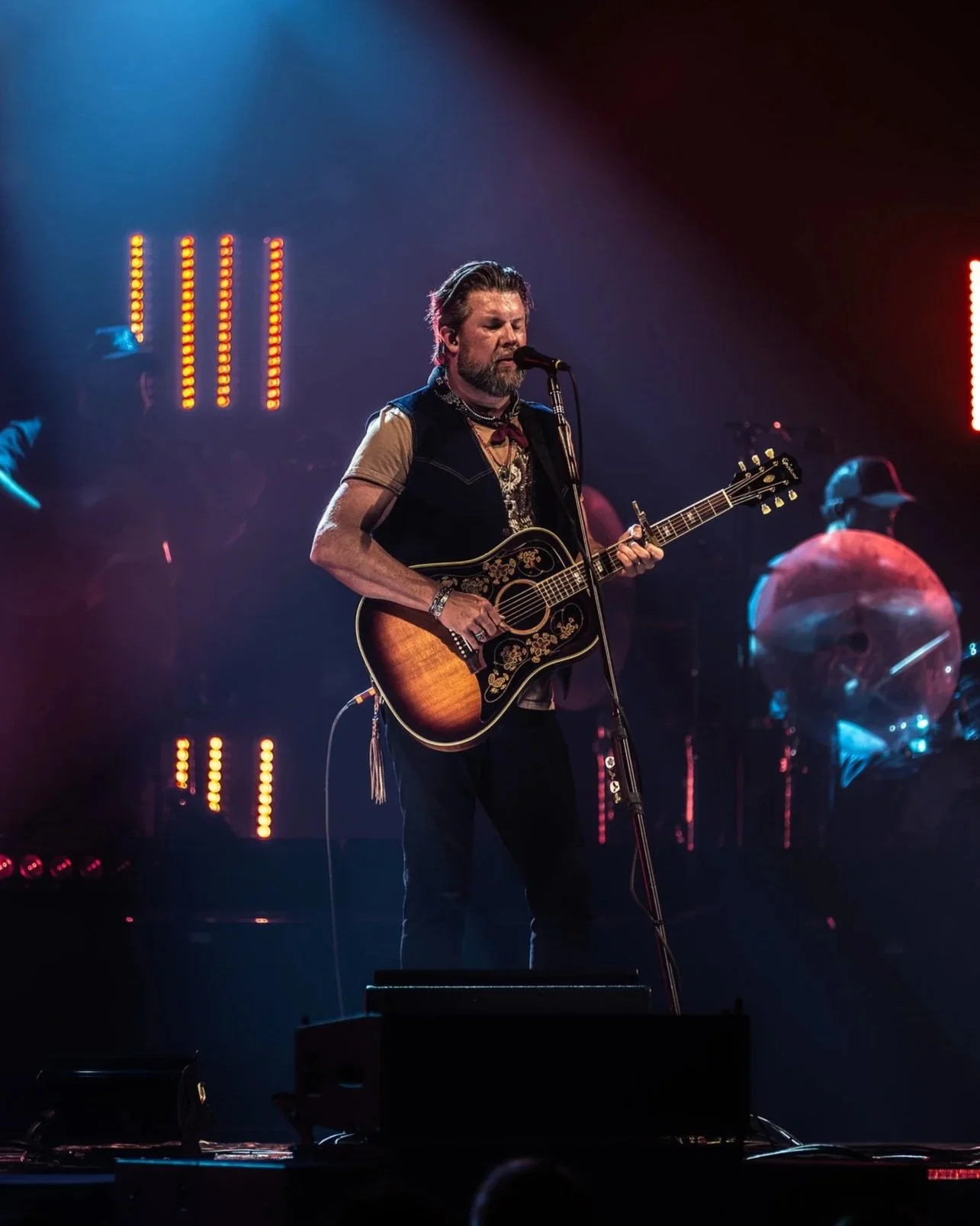 A male musician with a beard and styled hair, performing on stage with an acoustic guitar. He is singing into a microphone, dressed in a beige shirt with a black vest, and surrounded by colorful stage lights.
