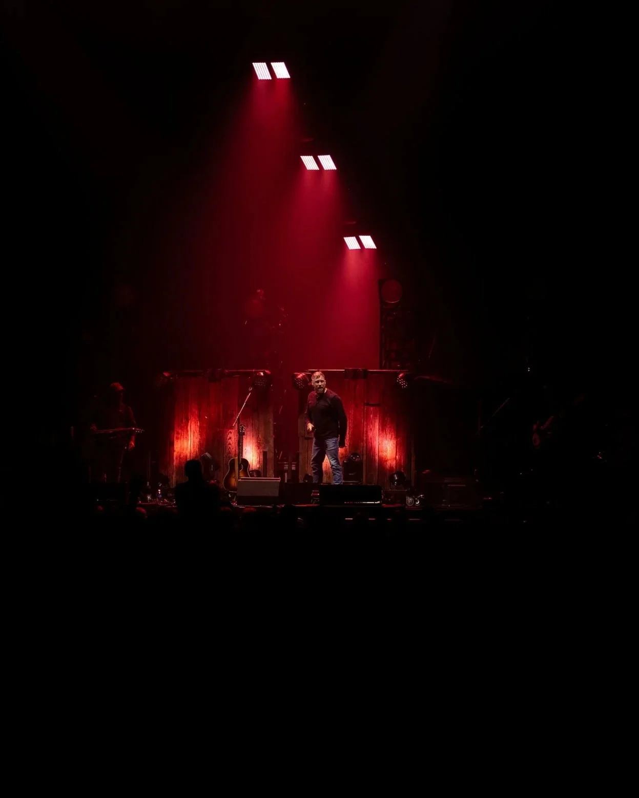 Performer on stage during a concert, surrounded by musical instruments and red light effects.