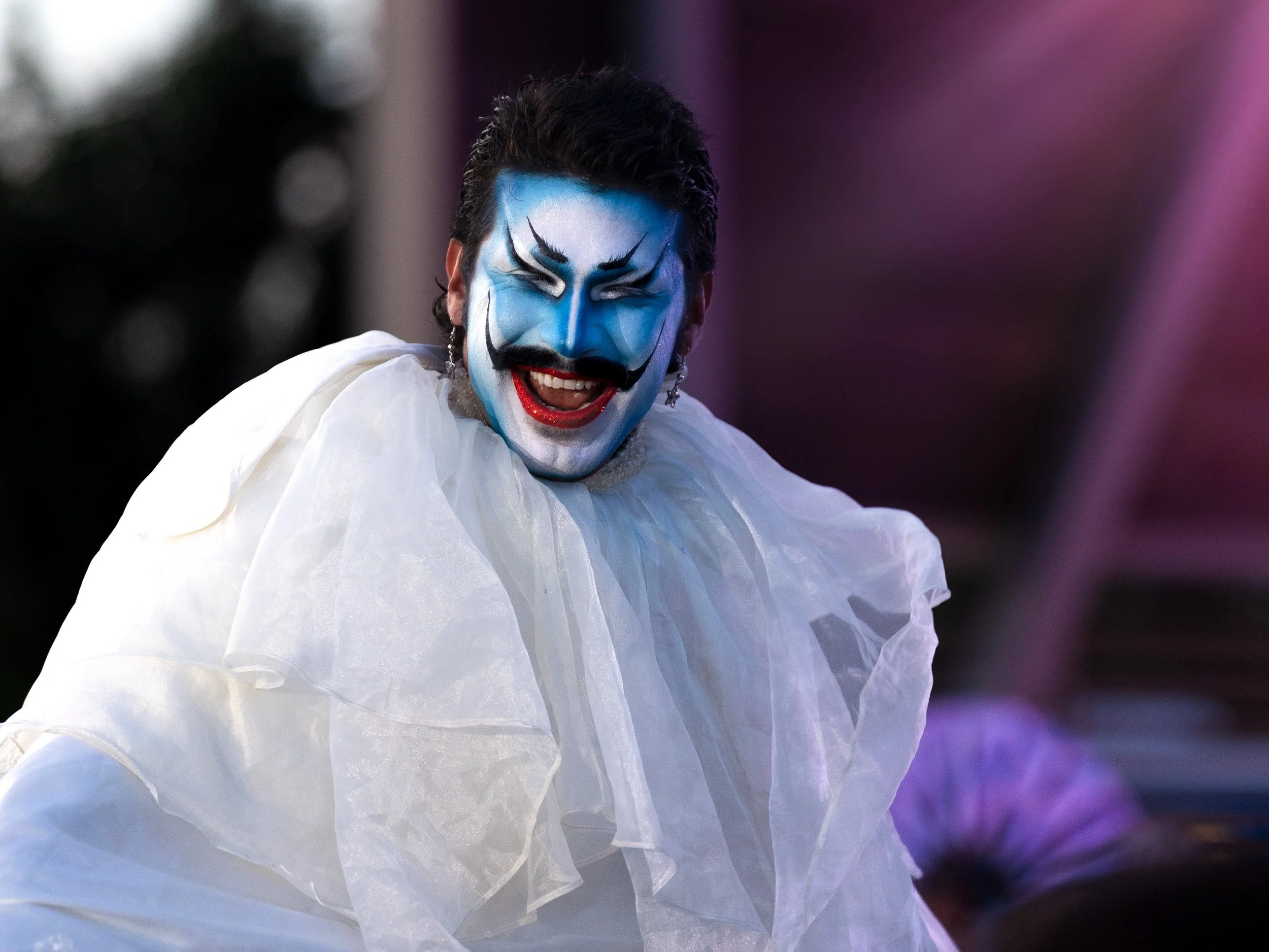 Person dressed in clown makeup with blue face paint, white makeup, and black facial features, wearing a white ruffled costume, smiling while performing outdoors.