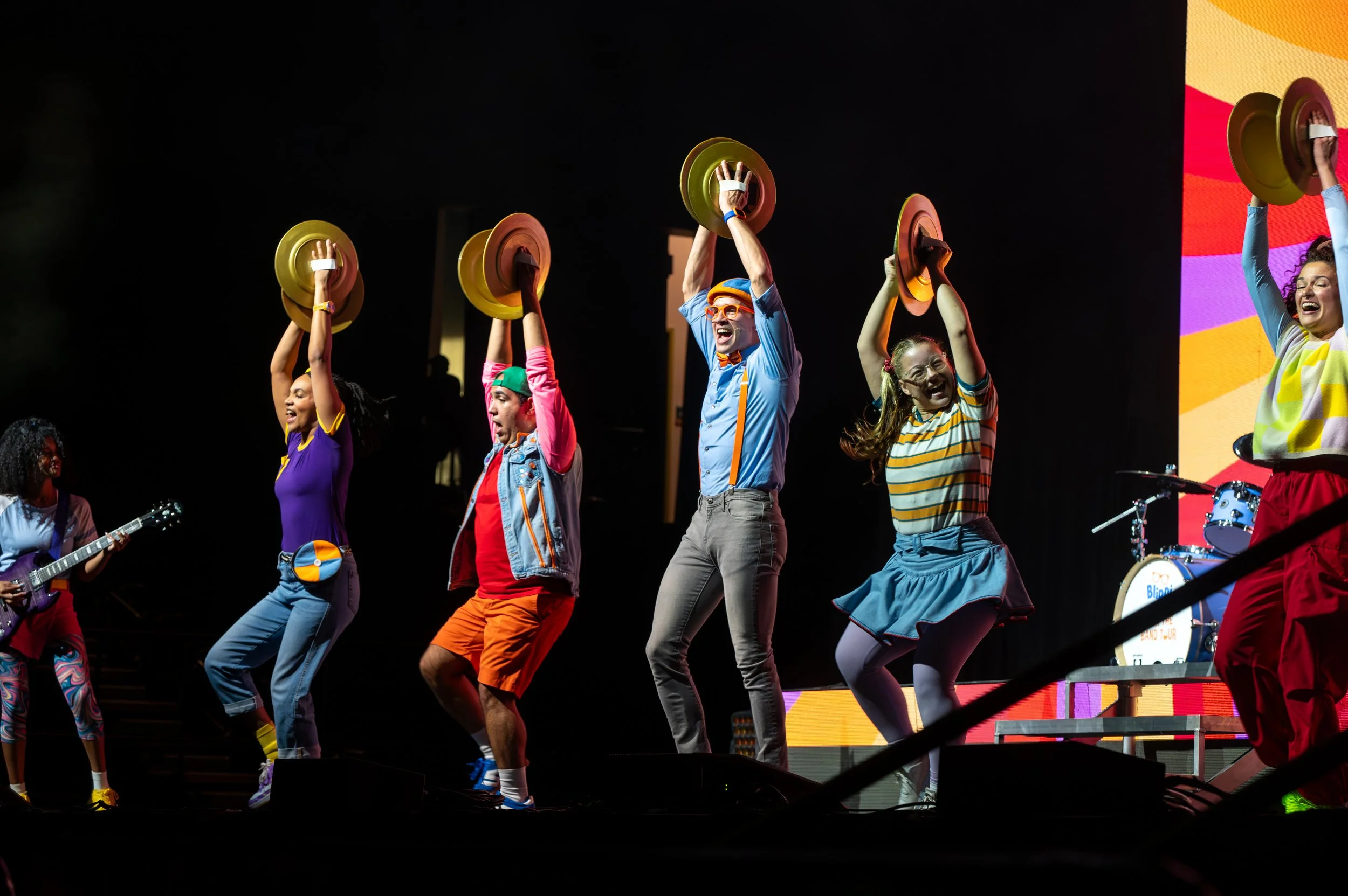 A group of performers on stage holding colorful bowls or pots above their heads during a lively performance with a musician playing guitar on the side and a colorful background.