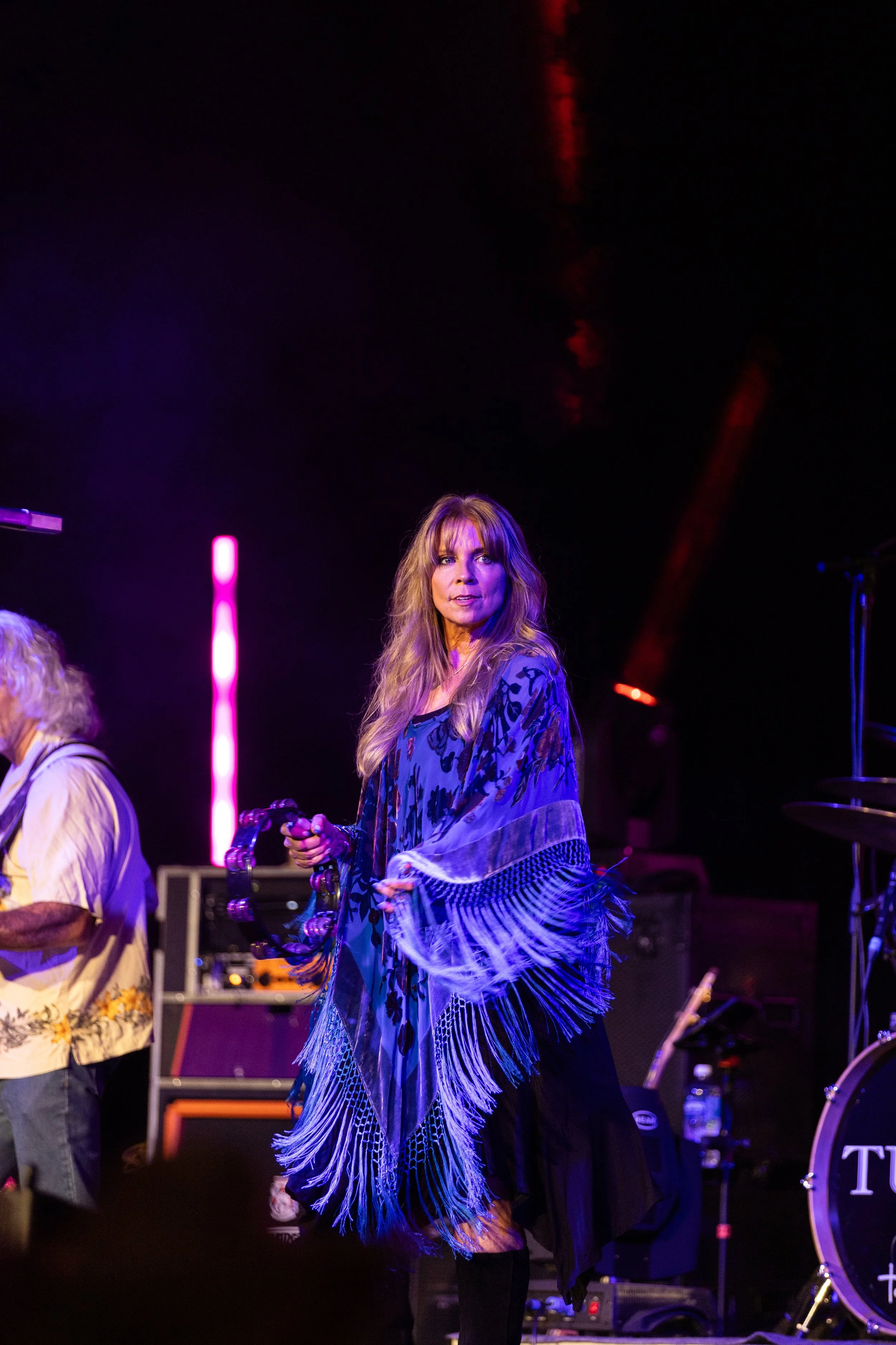 A woman with long blonde hair in a blue and black scarf performing on stage with purple and red lighting.
