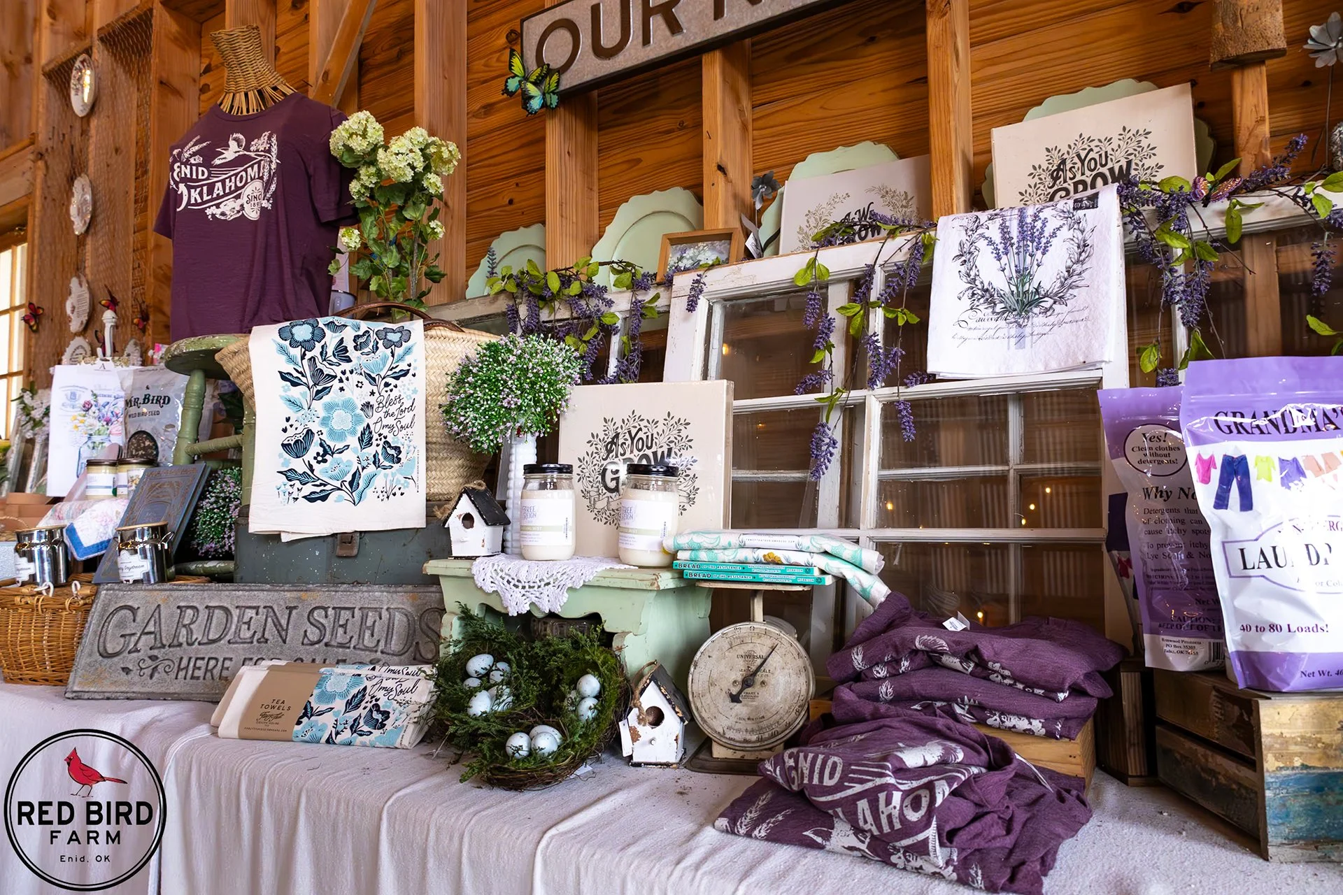 A market display at Red Bird Farm in Enid, Oklahoma, featuring gardening and home decor items, textiles, potted plants, and signs with floral and nature-themed designs, arranged on a table and shelves against a wooden wall.
