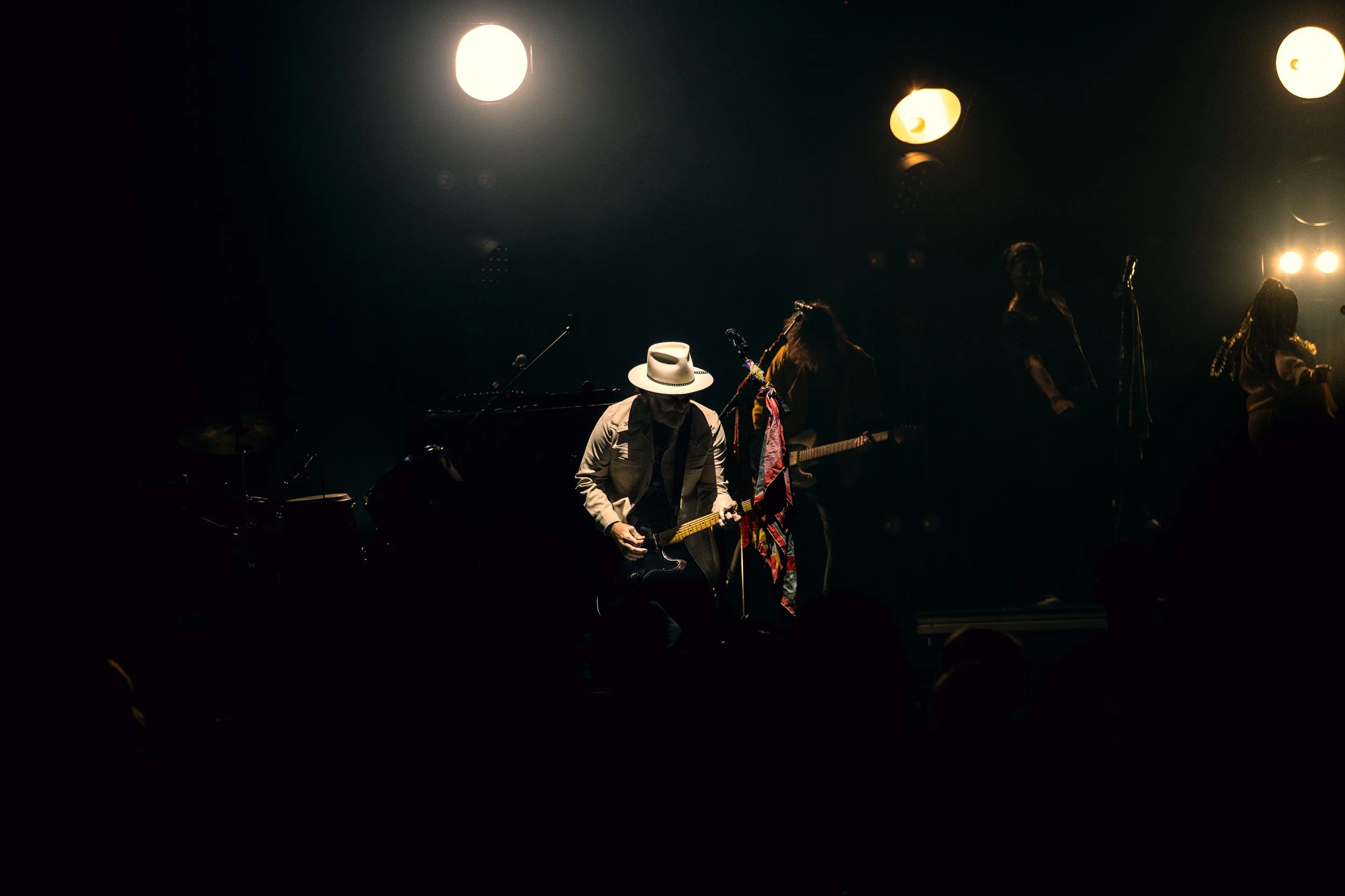Musicians performing on a dark stage with bright spotlights, including a guitarist wearing a white hat and a vest.