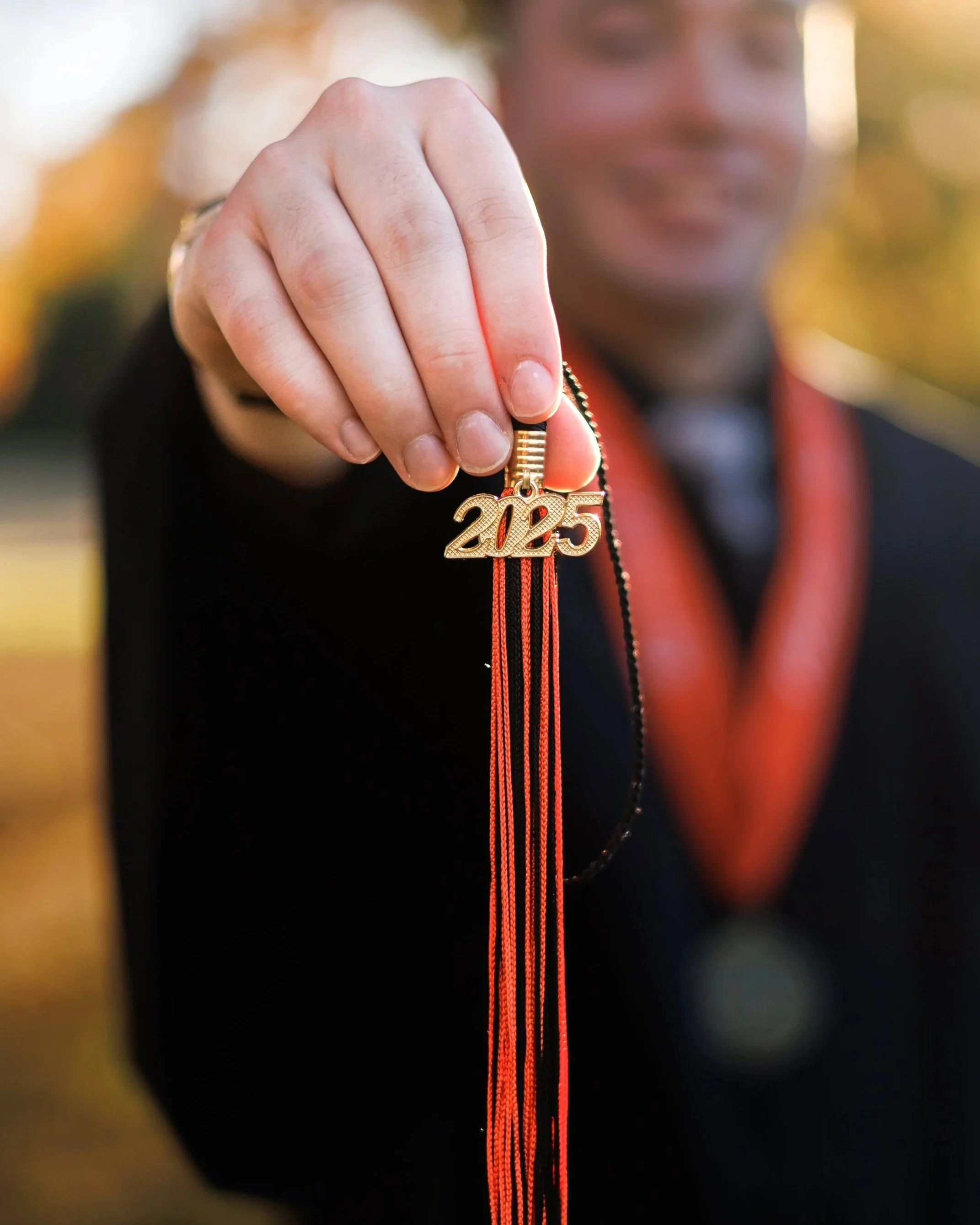 Person holding a gold graduation tassel with a '2023' charm, wearing a medal around their neck, celebrating graduation outdoors.